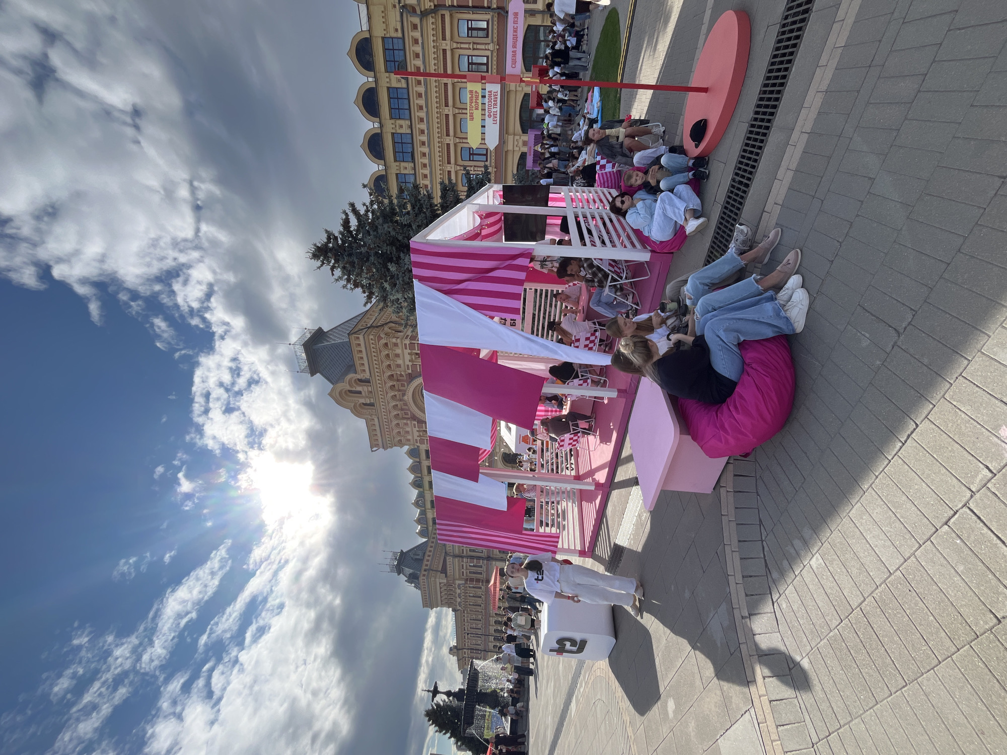 In Nizhny Novgorod, Russia, on a sunny day with scattered clouds, people are gathered in a public square. A pink and white striped tent, likely a promotional setup, is set up in the center of the scene. Several people are seated on beanbag chairs and around small tables within and around the tent, some appear to be engaging in conversation. A young woman in white stands near a large white cube with a logo. In the background, a large, ornate building, possibly a historical landmark, stands with many windows. The ground is paved with rectangular gray bricks. There is a fountain in the distance to the left, and a large tree to the right of the tent. Signage in Russian is visible on the building in the background.