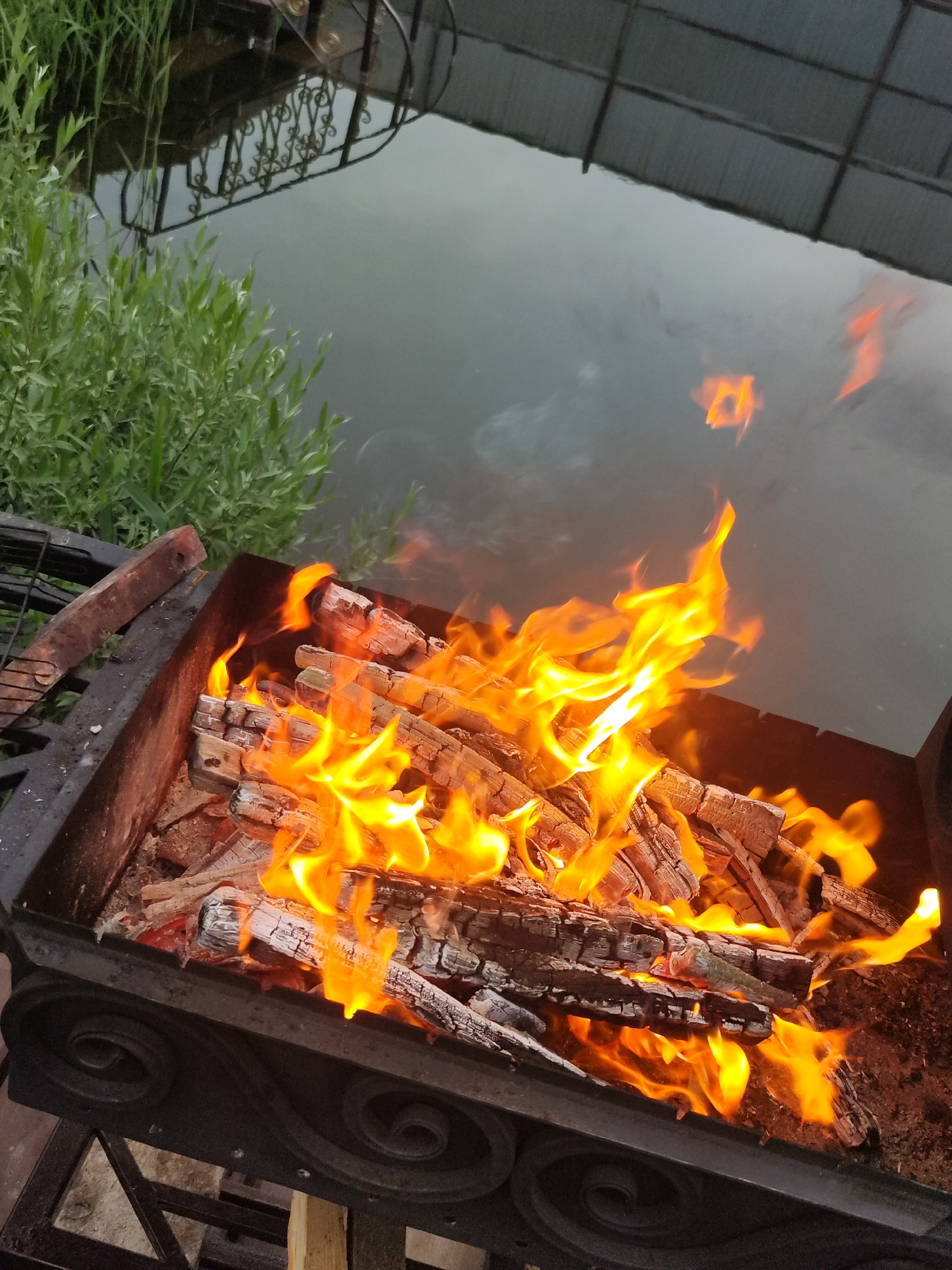 The image depicts a metal grill filled with burning wood, producing vibrant flames. In the background, slightly out of focus, is a body of water, possibly a river or lake, reflecting the sky and nearby structures. To the left of the water, green foliage is visible. The location appears to be Chaikovsky, Russia.
