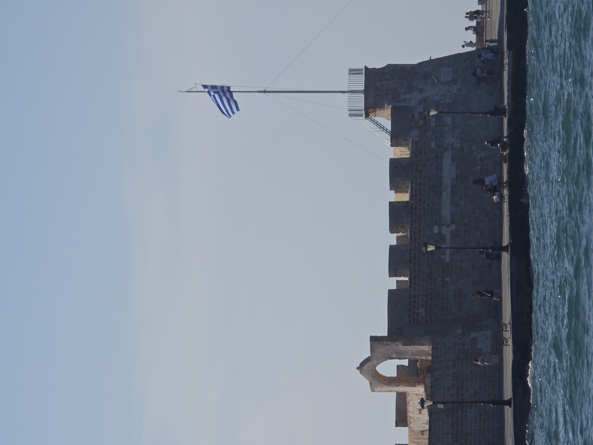 The image depicts the Venetian harbor of Chania, Greece. A stone fortress wall, weathered and aged, dominates the mid-ground. The wall features an arched opening on the left and battlements along its top. A tall flagpole rises from a higher section of the fortress, flying the blue and white Greek flag. People are visible walking along the walkway in front of the fortress, some sitting on benches. Two lampposts line the walkway. The foreground shows the blue, choppy waters of the harbor. The sky is a pale blue with a few faint clouds. The scene is bathed in bright sunlight, casting clear shadows.