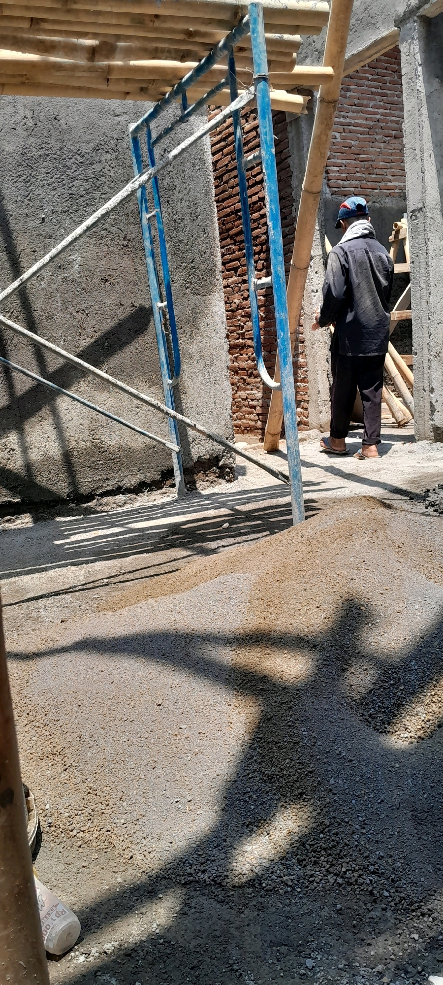The image depicts a construction site in Karangduren, Indonesia. A man, wearing a dark long-sleeved shirt, dark pants, a cap, and sandals, stands with his back to the camera. He appears to be holding a tool or object in his right hand. The man is standing near a pile of aggregate or gravel, and scaffolding structures are visible throughout the scene, constructed with blue metal poles and bamboo. A partially constructed brick wall is present in the background. The ground is uneven and covered with gravel and dirt. Shadows cast by the scaffolding and possibly the man are prominent on the ground. The overall environment suggests an active construction zone.