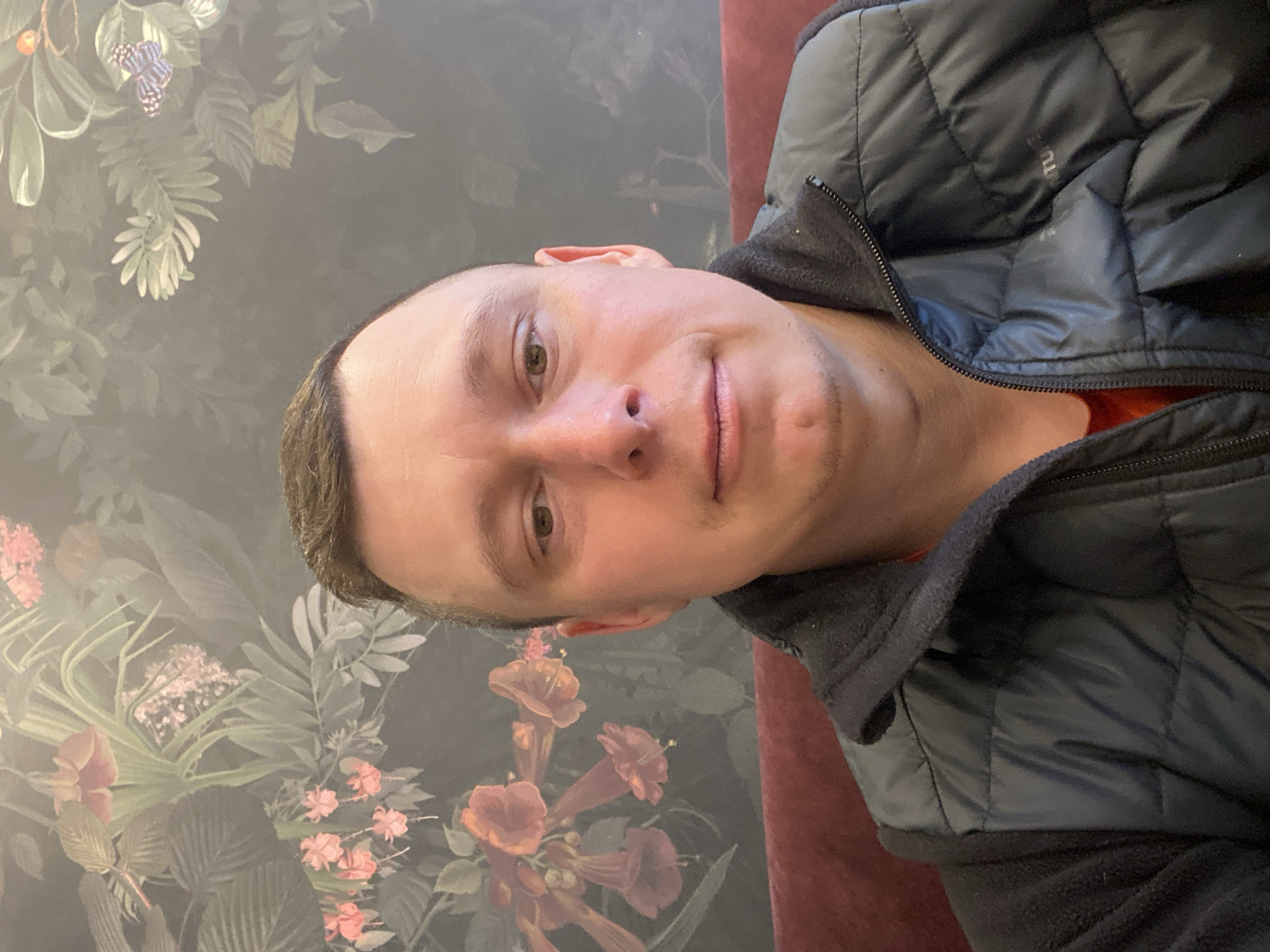A man wearing a black jacket is in front of a floral patterned wall. The man has short hair and light skin. The location is not identifiable as Neftekamsk, Russia, without additional context or information.