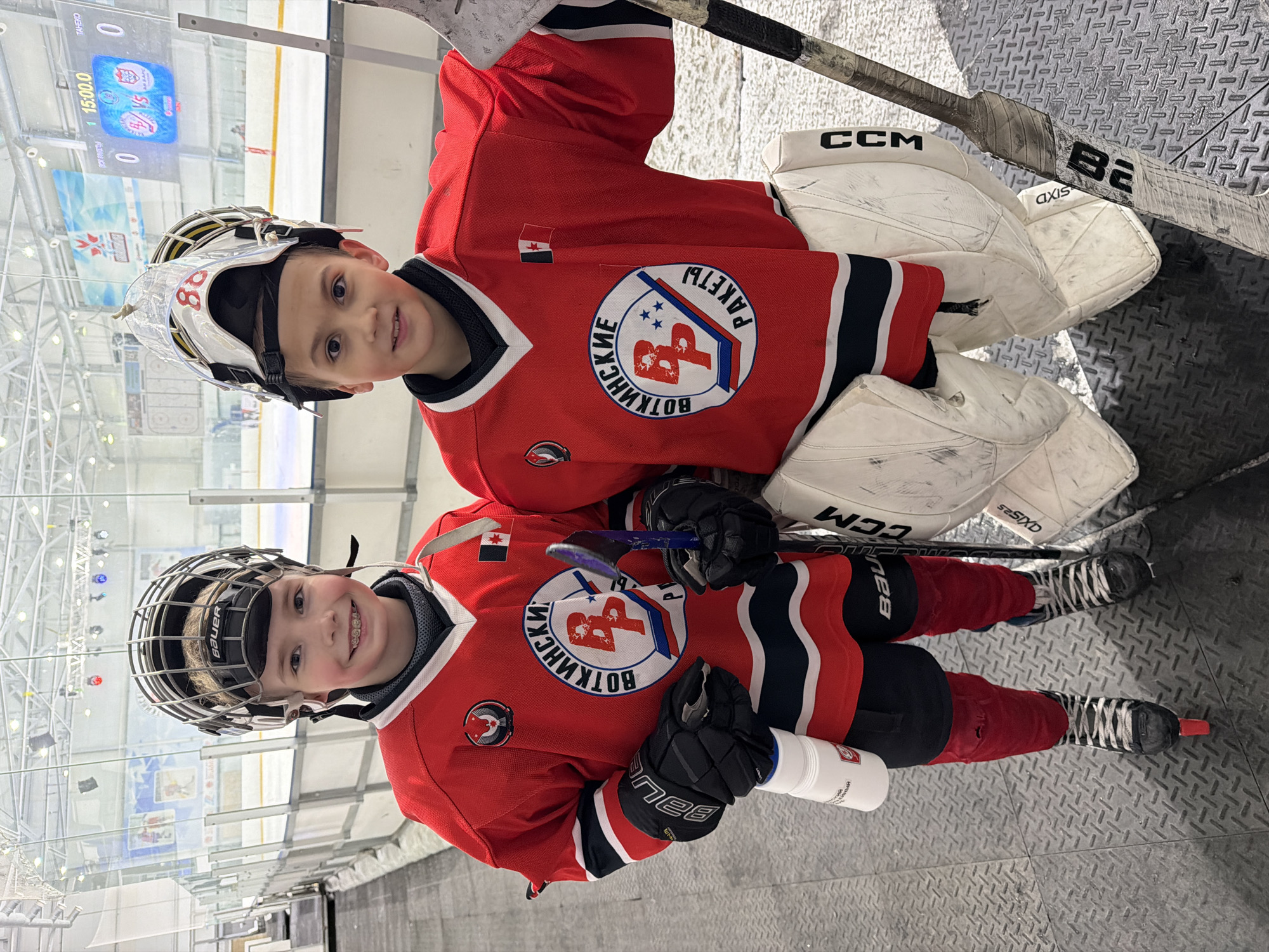 Two young boys wearing red and black hockey jerseys with the logo "ВОТКИНСКИЕ РАКЕТЫ" (Votkinsk Rockets) are posing for a photo. The boys are wearing hockey helmets, with one wearing a goalie mask. The boys are holding hockey sticks and wearing hockey gloves. The background shows an ice rink with glass barriers and a scoreboard showing a time of 15:00.0. The logo on the jerseys and the name "ВОТКИНСКИЕ" indicate the location is likely Votkinsk, Russia.