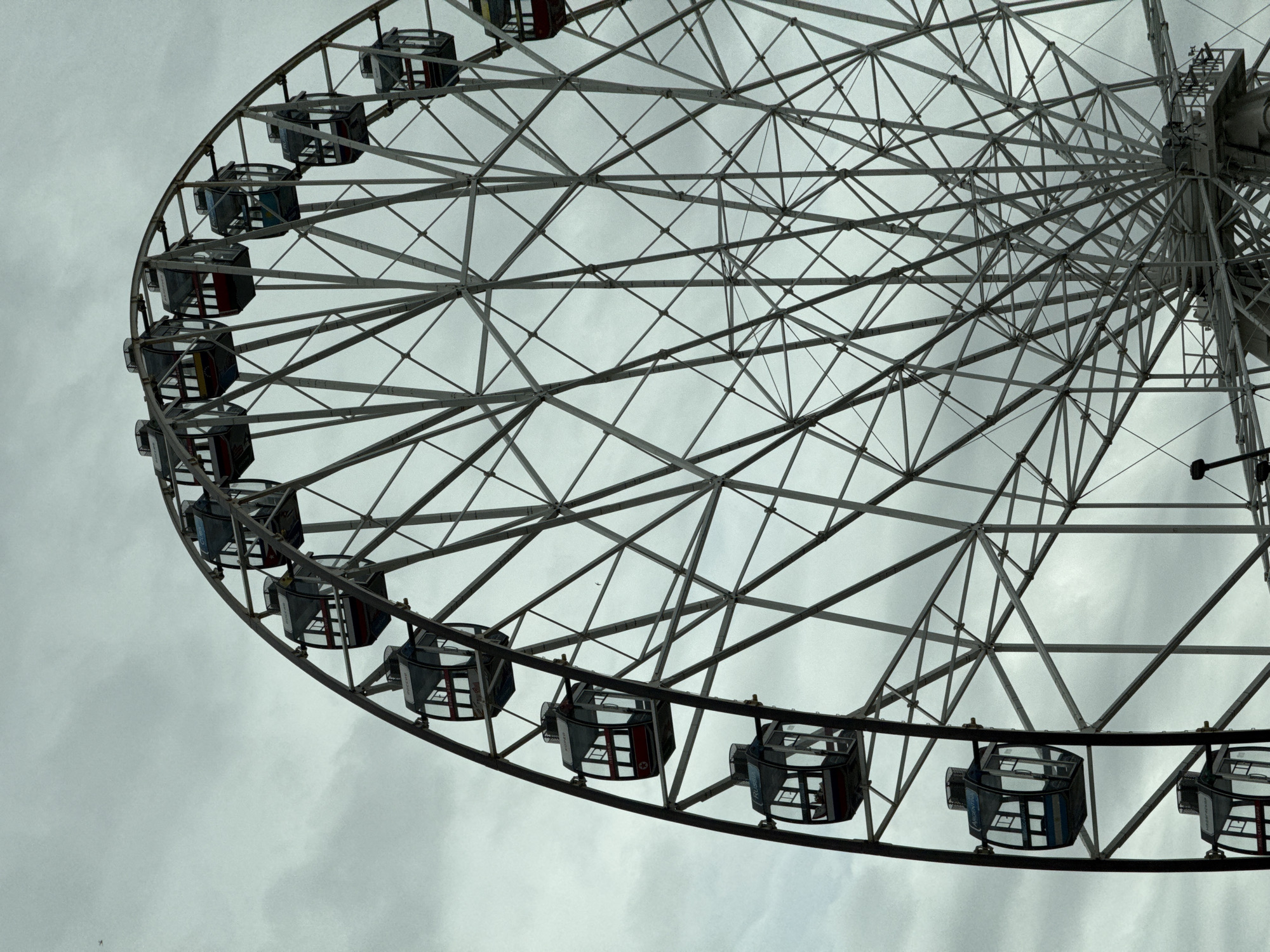 The image depicts a portion of a Ferris wheel against a cloudy sky. The wheel's structure consists of a network of gray metal beams and support frames. Passenger cars are visible, appearing dark and boxy with glass windows. The image is taken from a low angle, looking up at the wheel. The photograph was taken in Kazan, Russia.
