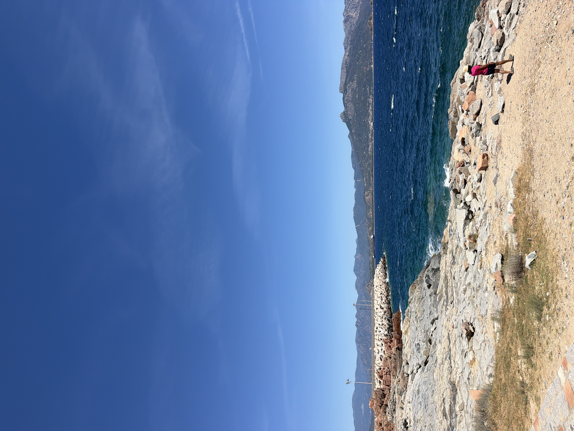 The image depicts a coastal scene in Tortolì, Italy, characterized by a rocky shoreline and the deep blue Mediterranean Sea under a clear sky. In the foreground, a person wearing a pink t-shirt and dark shorts walks on a sandy and rocky path. The path is strewn with numerous rocks of various sizes and colors, with patches of dry grass growing between them.
The middle ground features the vibrant blue sea with moderate waves lapping against the shore. In the distance, a breakwater made of concrete blocks extends into the water, with lampposts visible on it. Beyond the breakwater, a hilly coastline with sparse vegetation rises against the horizon. Further in the distance, a ship is visible on the water. The background is dominated by a vast expanse of clear, bright blue sky with a few wispy white clouds. The overall atmosphere is one of a sunny, outdoor excursion by the sea.