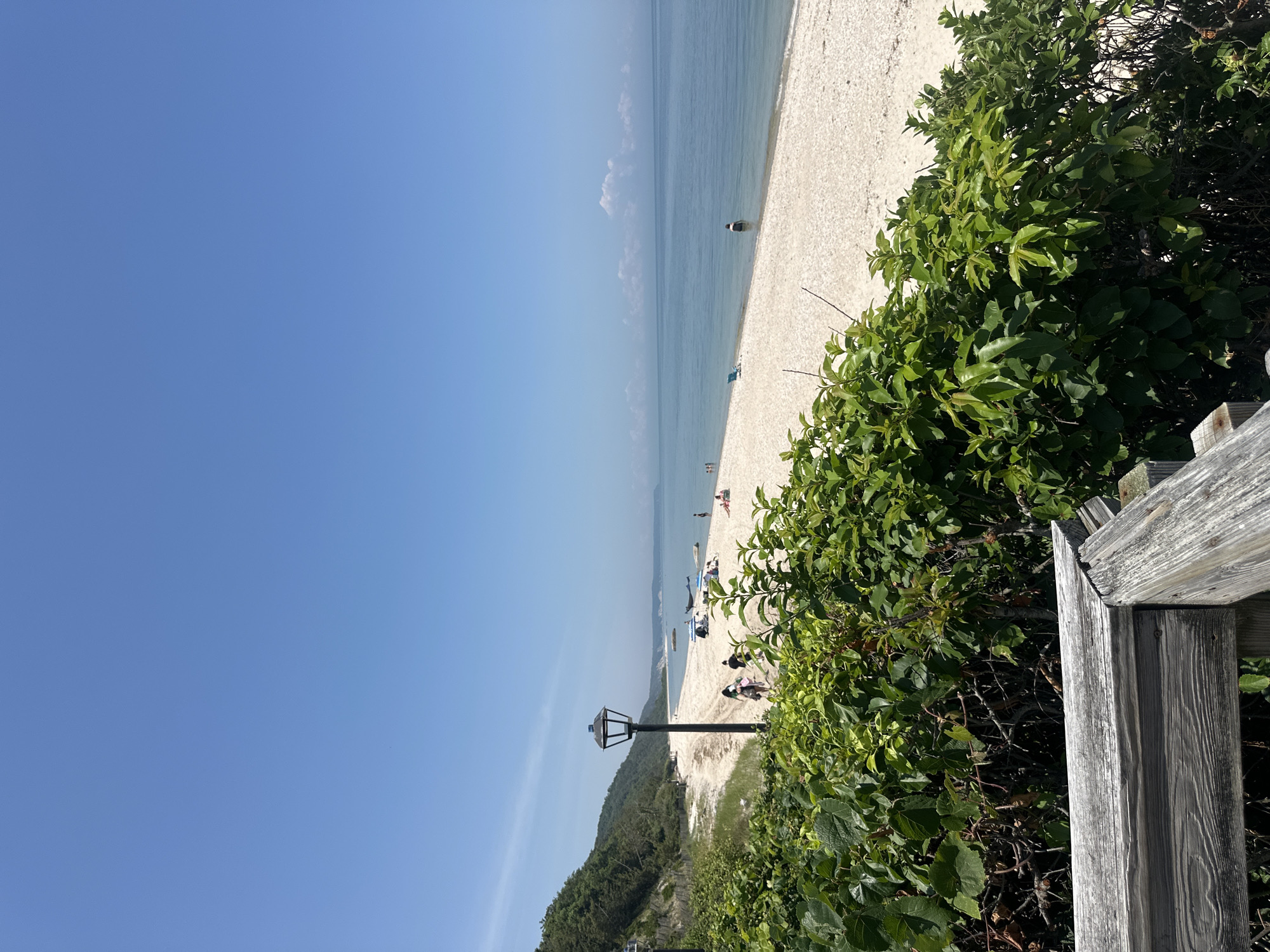 The image shows a sunny day at a beach, likely in the Town of Riverhead, United States of America. In the foreground, a wooden railing and lush green foliage obscure the lower part of the scene. Beyond the vegetation, a paved walkway with a lamppost runs parallel to a sandy and pebbly beach. Several people are visible on the beach, some walking, others sitting or standing. A few beach umbrellas and chairs are scattered across the sand, suggesting recreational activity. The ocean stretches out to the horizon, with calm, blue water. In the distance, a tree-covered hillside forms the backdrop to the beach. The sky is clear and a bright blue, with a few wispy clouds.