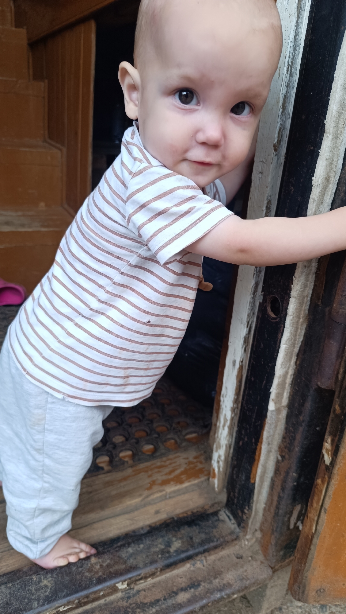 A very young Caucasian child is standing and leaning on a wooden doorway. The child has fair skin and is wearing a light-colored t-shirt with thin brown stripes and light grey pants. The child is facing the camera and looking directly at the viewer with a slight smile. The background shows wooden stairs on the left and a dark interior on the right. The doorway itself appears weathered and aged, with peeling paint and visible hardware. The floor in the doorway has a textured mat. The scene is likely outdoors, near the entrance to a building in Karsha, Russia.