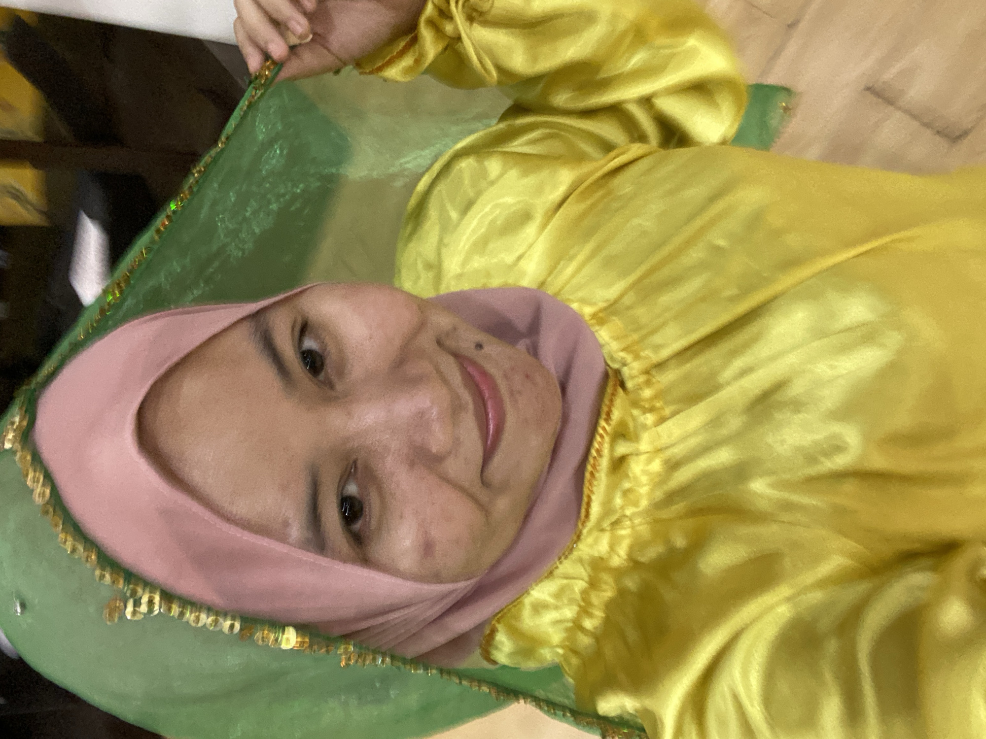 The image shows a close-up of a woman wearing a pink hijab and a shiny yellow garment. A green veil with gold trim is draped over her head and shoulder, and she is holding it up with her right hand. The woman is smiling and looking at the camera. The background is out of focus, but appears to be indoors with wooden flooring and hints of yellow and dark colors. There are no explicit visual cues in the image to determine a specific location within Banjarmasin, Indonesia.