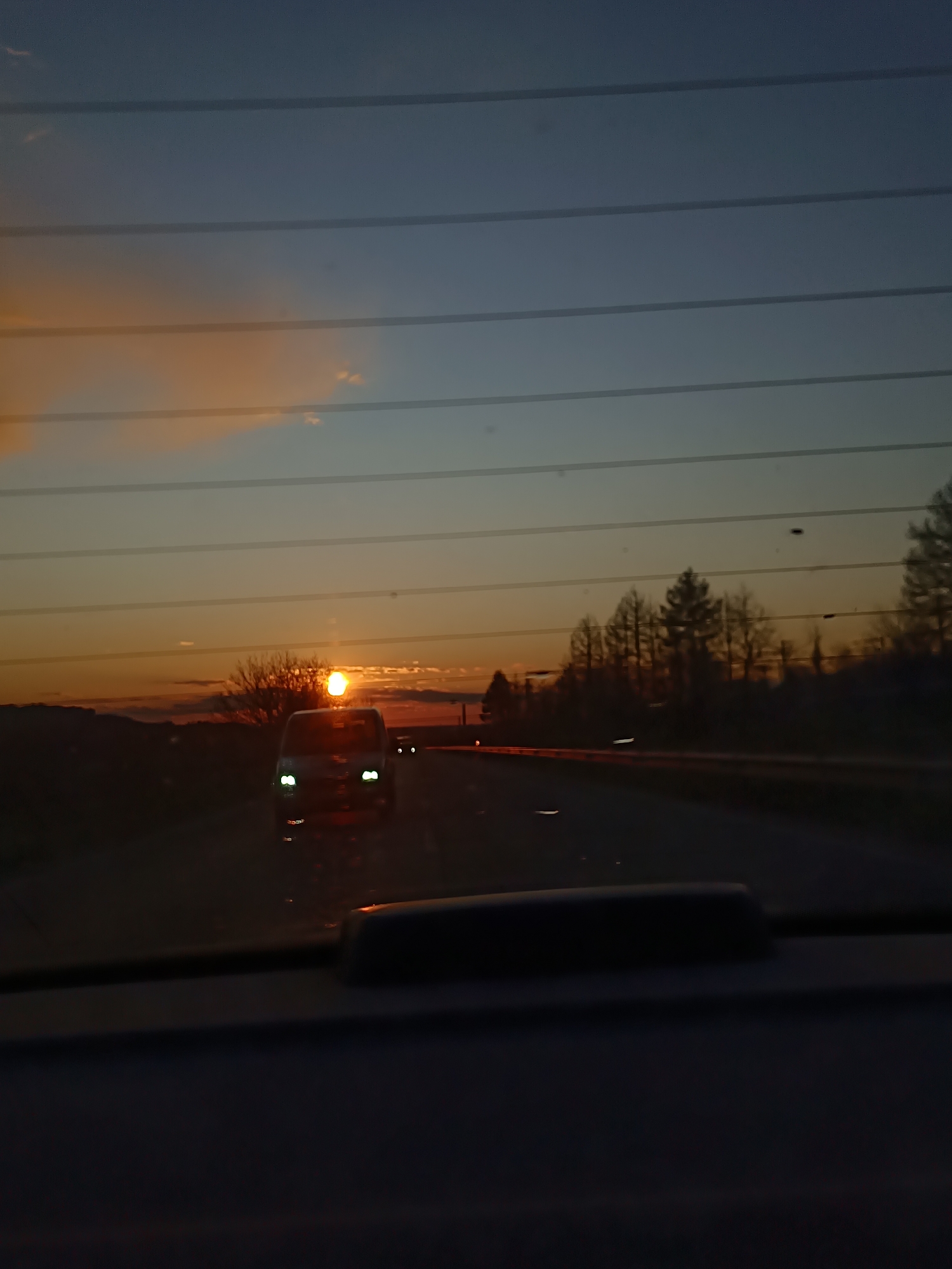 The image depicts a sunset viewed through the rear window of a vehicle. The sky displays hues of orange and blue, bisected by parallel horizontal lines, likely defroster wires. In the distance, a car is traveling on the road with its headlights illuminated, appearing brighter due to the sun's position. Silhouetted trees line the horizon, creating a dark contrast against the setting sun. The bottom portion of the image features the interior of the vehicle, including the rear deck and headrests. The location appears to be a highway or road outside a city, judging by the sparse development and presence of trees. The location is given as Unknown, Unknown.