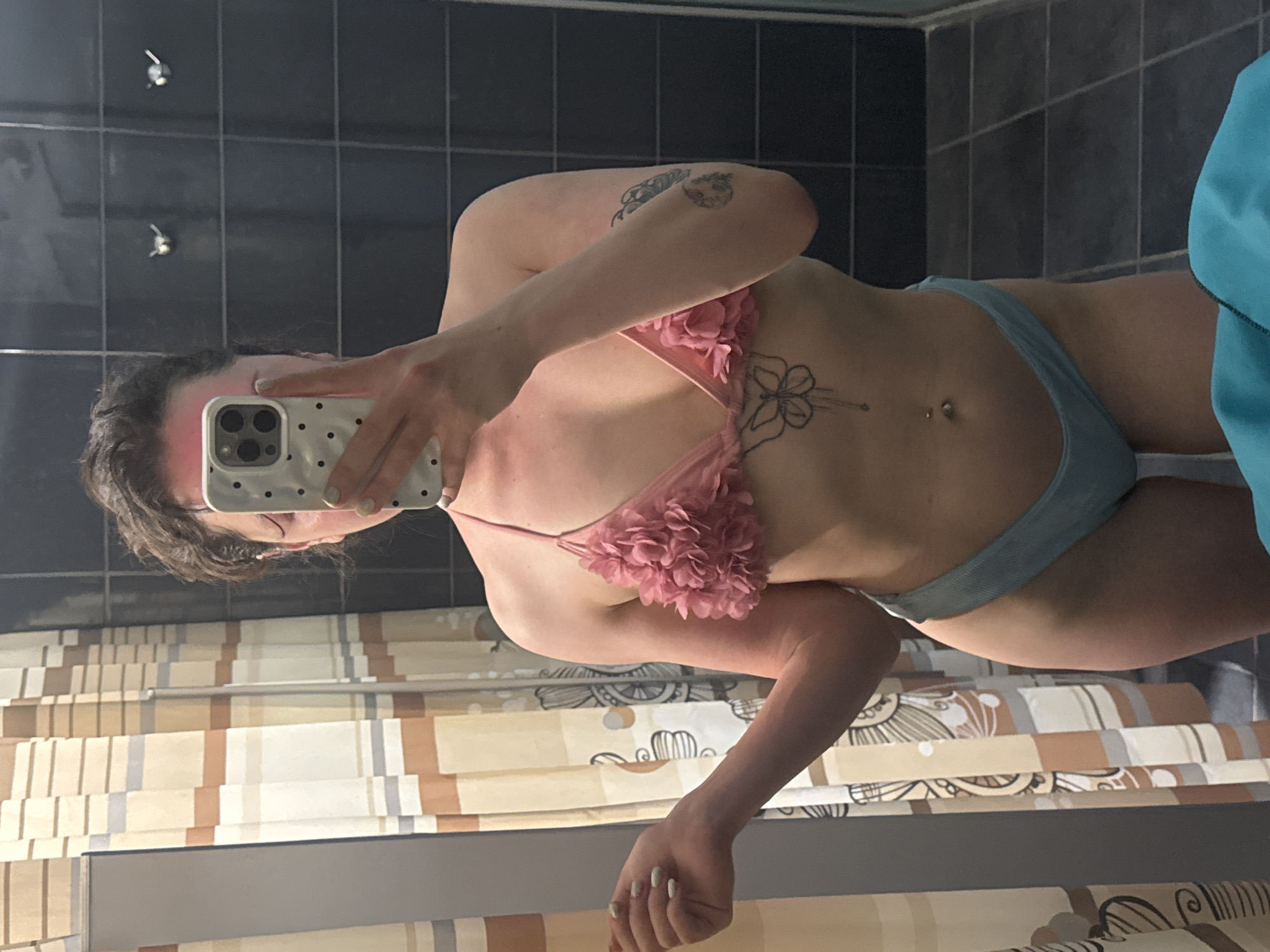 A person is captured in a mirror selfie, standing in what appears to be a bathroom. The subject is wearing a two-piece swimsuit: a pink bikini top adorned with floral ruffles and light blue bikini bottoms. Their right arm is raised, holding a white smartphone with a black polka-dot case and a triple camera system, which partially obscures their face. The person's hair is dark and appears to be styled loosely or pulled back. A reddish tint is visible on their forehead, possibly due to lighting or a tan.
Visible tattoos include a cartoon-style character on their upper right arm/shoulder, and a floral design with a stem extending towards the navel area on their sternum/upper abdomen. A belly button piercing is also visible. The person's left hand, with light-colored nail polish, rests casually by their side.
The background reflects a dark grey or black tiled wall with white grout lines. Two small, metallic hooks are visible on the tiled surface. Below the tiled wall, a shower curtain with vertical stripes in shades of beige, brown, and light grey, along with abstract circular or floral patterns, is visible. A sliver of blue fabric, possibly a towel, is seen on the far right. The setting is consistent with a private bathroom, likely in Burgas, Bulgaria, given the provided location context. There is no visible text in the image.
