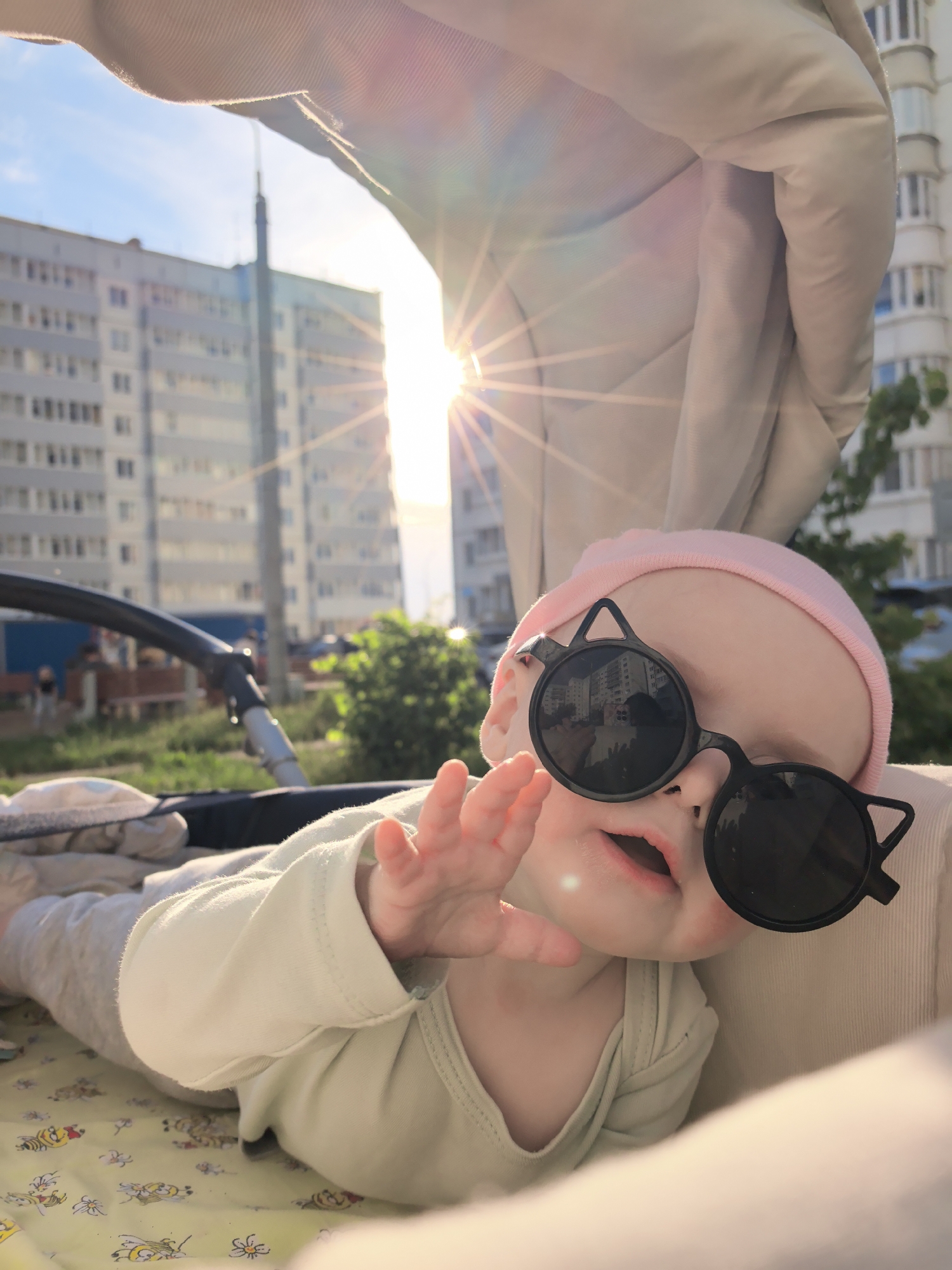 A baby, wearing a pink hat and cat-eye sunglasses, lies in a baby stroller. The baby is wearing a light green onesie and extending their left hand. The stroller has a tan colored canopy and the patterned stroller liner has bees on it. Behind the baby and stroller, apartment buildings are visible in the background, suggesting an urban setting. The sun is shining brightly behind the buildings, causing lens flare. The city is Фролы, Russia.