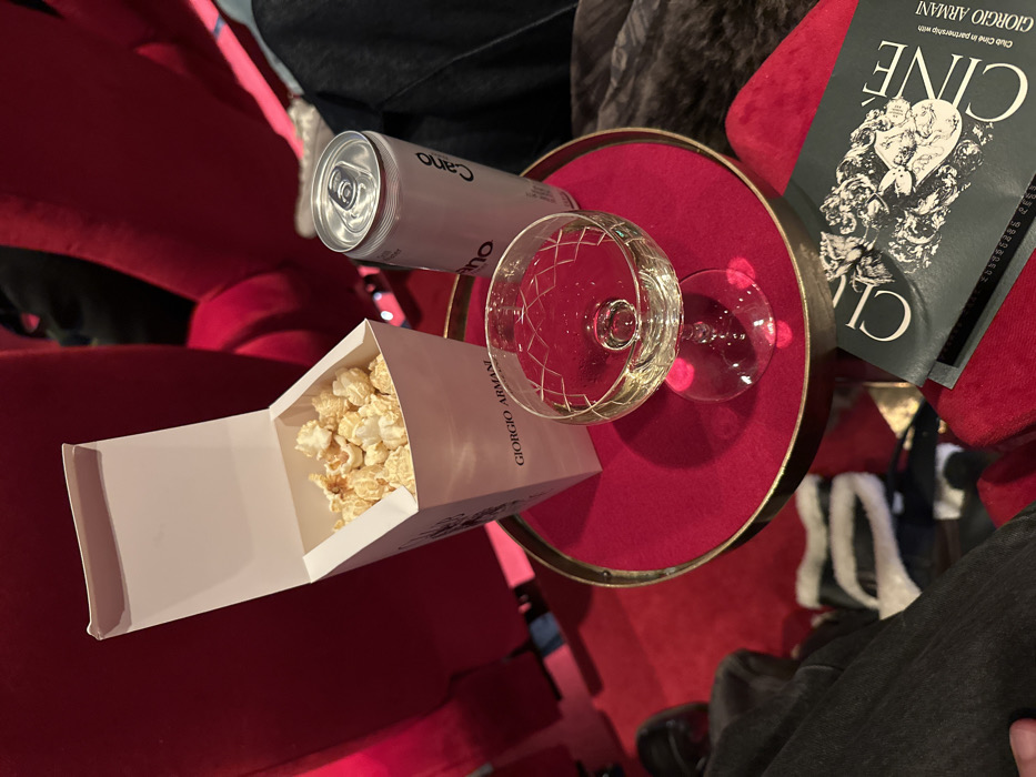 A round, red-velvet table holds a crystal glass of white wine, a silver can, and a white box of popcorn, all slightly tilted. A black and white Giorgio Armani brochure sits atop the table. The table is surrounded by red seating. Someone in black jeans and shoes sits to the side. The overall scene suggests a luxurious and indulgent movie-going experience.