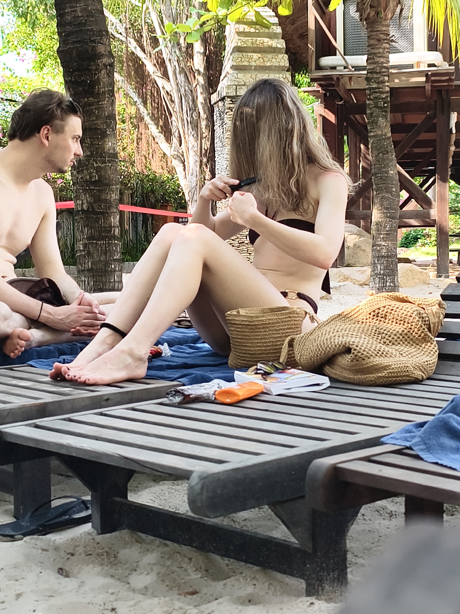 A medium shot captures a woman sitting on a grey wooden beach lounge chair, facing right and combing her hair. She is wearing a dark brown bikini. Next to her is a woven beach bag. A man sits on the lounge chair to her left, facing the woman. The photo was likely taken outside, as trees and what appears to be an elevated wooden structure are visible in the background. A blue towel is draped on the beach lounge chair beneath the woman. The location is Unknown, Unknown.