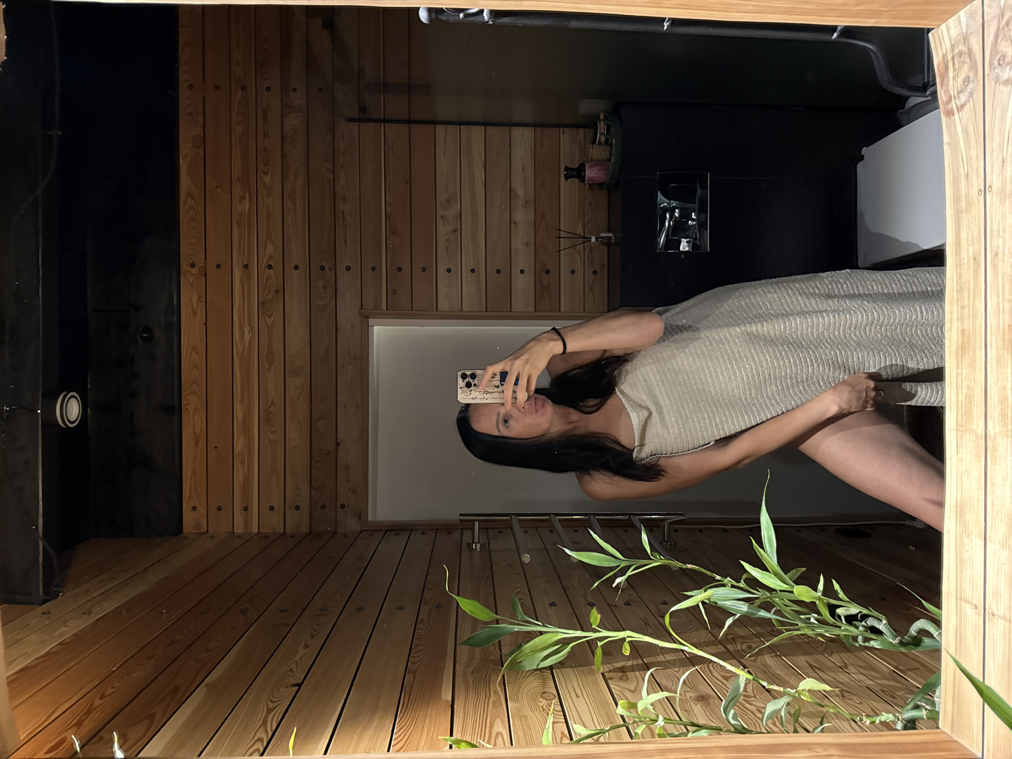 The image shows a woman taking a mirror selfie in a bathroom with wooden walls. She is wearing a towel wrapped around her body and is holding a smartphone. In the foreground, there are green plants. In the background, there is a dark cabinet with some bottles and diffusers. The location is specified as Chaikovsky, Russia.