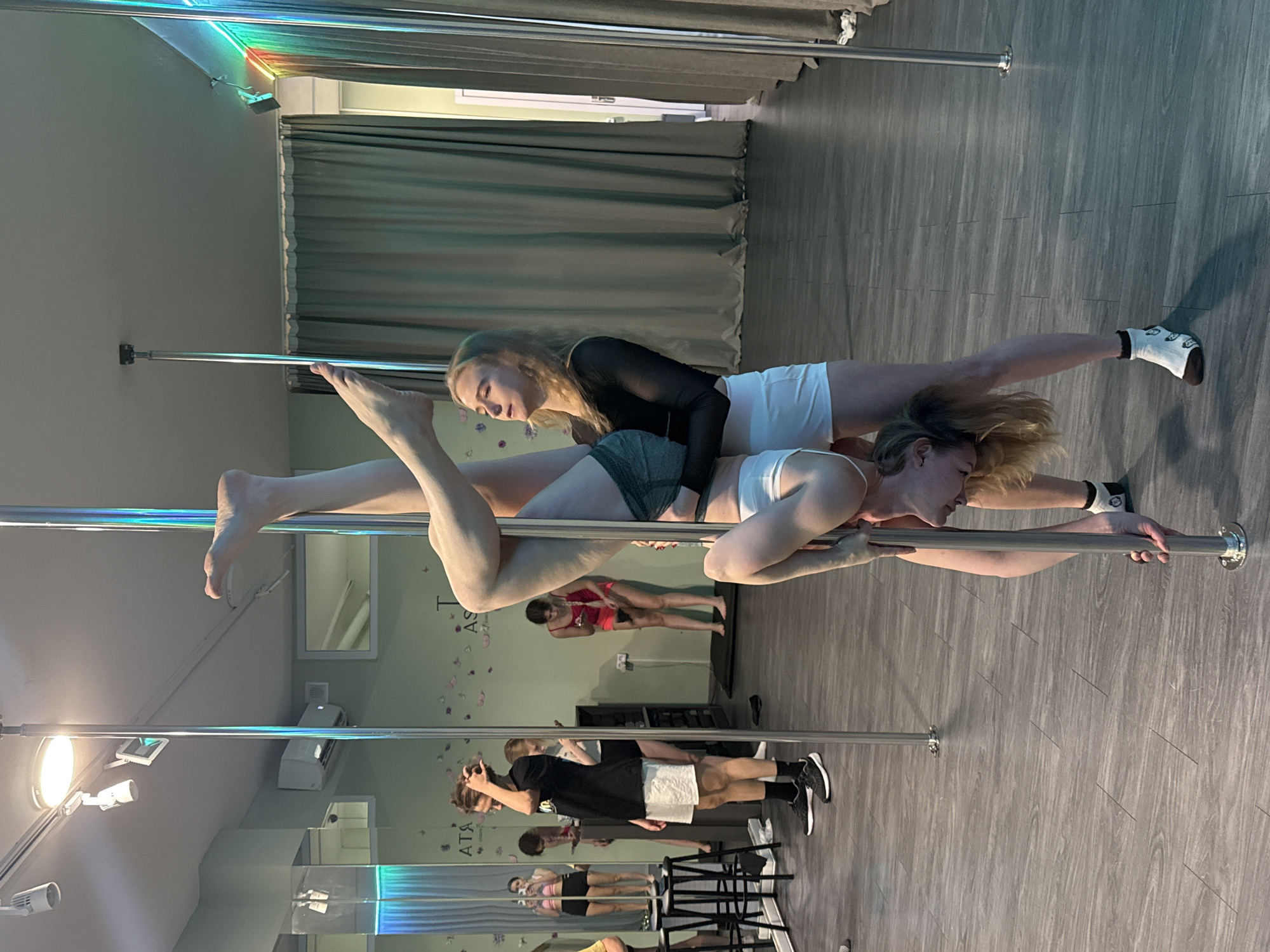 Two women are performing pole dancing in a studio setting. One woman, wearing a white top and shorts, supports the second woman, who is wearing a black long-sleeved shirt and dark shorts. The second woman is lying across the first woman's back, with her legs extended. Other individuals and poles are visible in the background, along with a mirror and a window with curtains. The environment appears to be a dance or fitness studio. The location is assumed to be in Batumi, Georgia.