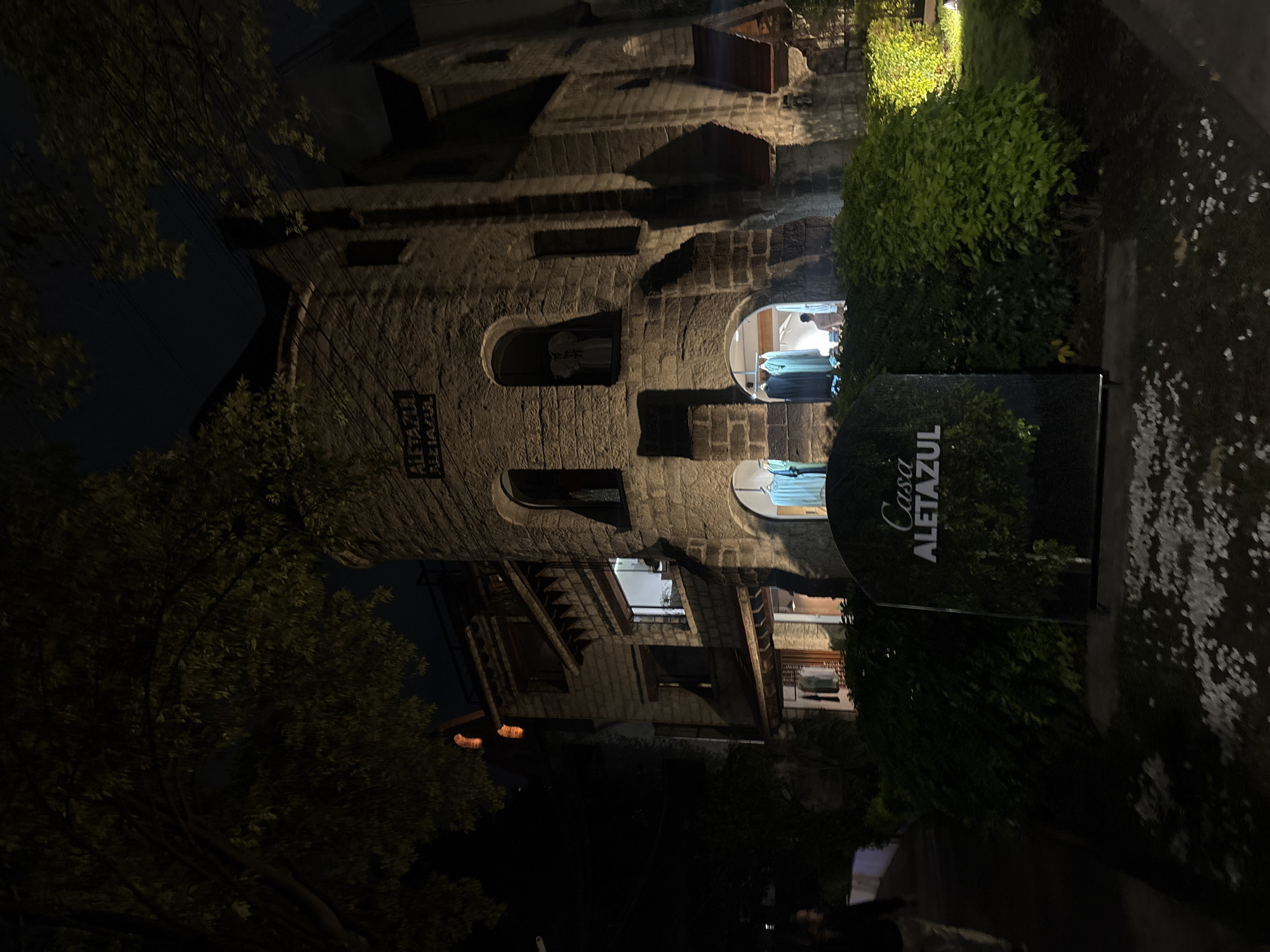 The image depicts the Casa Aletazul in Mexico City, Mexico, at night. The building is a tall, stone structure with arched windows. A sign reading "Casa Aletazul" is visible in front of the building. The scene is illuminated by artificial light. There is some vegetation next to the building, and a tree is visible to the left.