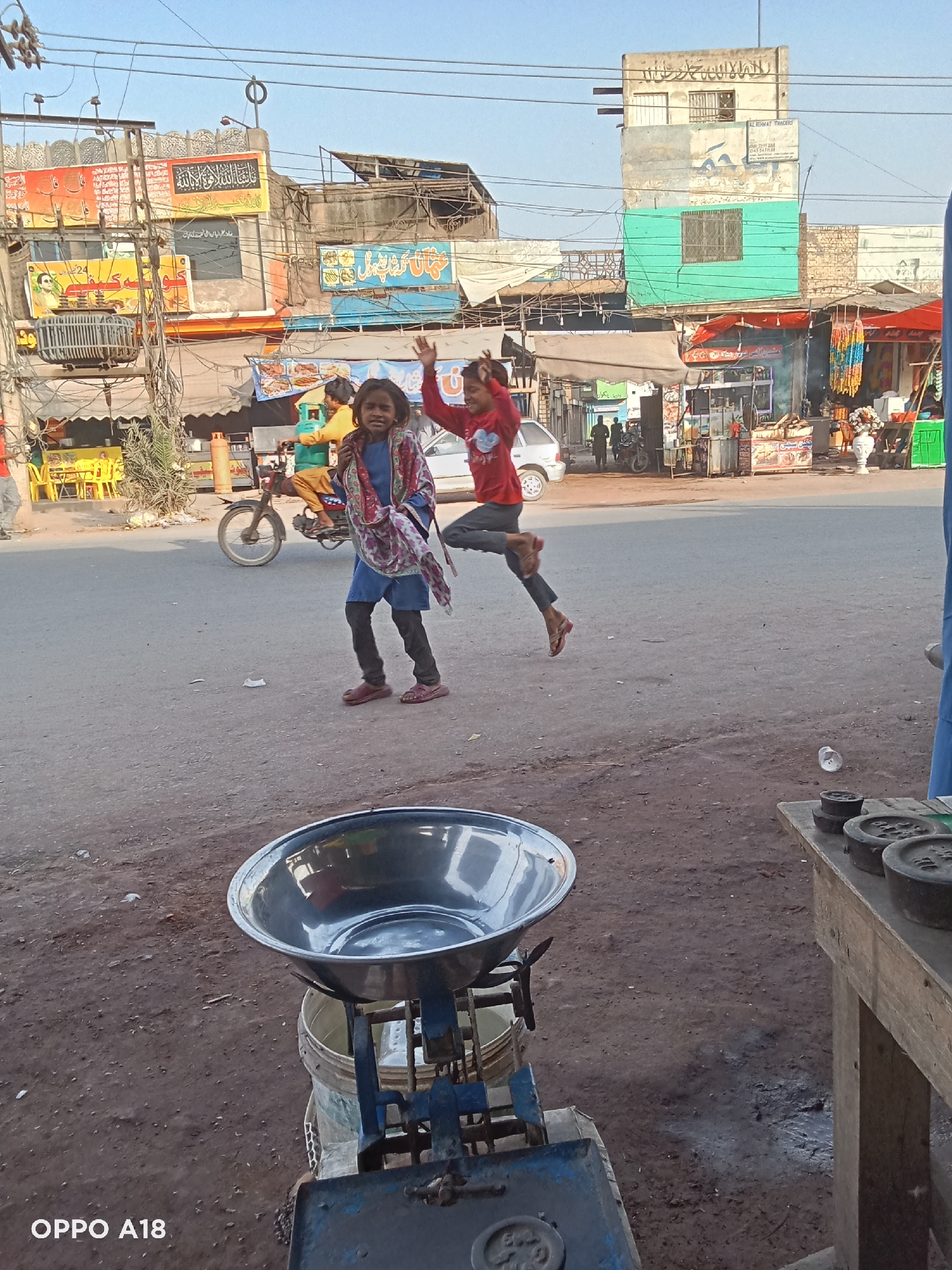 The image captures a vibrant street scene, likely in South Asia, during daytime under a clear sky. In the foreground, two young girls are the central subjects. One girl, on the left, looks towards the camera with a neutral expression, wearing a blue tunic, dark pants, red sandals, and a colorful patterned shawl. Beside her, another girl is captured mid-air, jumping with a joyful expression and raised hands, dressed in a red long-sleeved shirt and dark pants.
In the immediate foreground, a large, metallic weighing scale with a bowl is partially visible on the left, next to a wooden table holding several heavy metal weights on the right. The ground is a mix of asphalt and dusty dirt.
The background reveals a bustling street lined with numerous shops and multi-storied buildings. These structures appear weathered and are adorned with various signs and banners. A person on a motorcycle, wearing yellow attire, can be seen in the middle ground, heading right. Overhead, a dense network of electrical wires crisscrosses between poles and buildings, including a prominent electrical transformer on the left.
Visible text includes an "OPPO A18" watermark at the bottom left. On the buildings, several signs feature Arabic or Urdu script. Notably, a large banner on a building on the left has prominent Arabic/Urdu calligraphy, and another sign reads "AL REHMAT TRADERS" with a phone number. The overall scene conveys a sense of everyday life and activity in an urban environment.