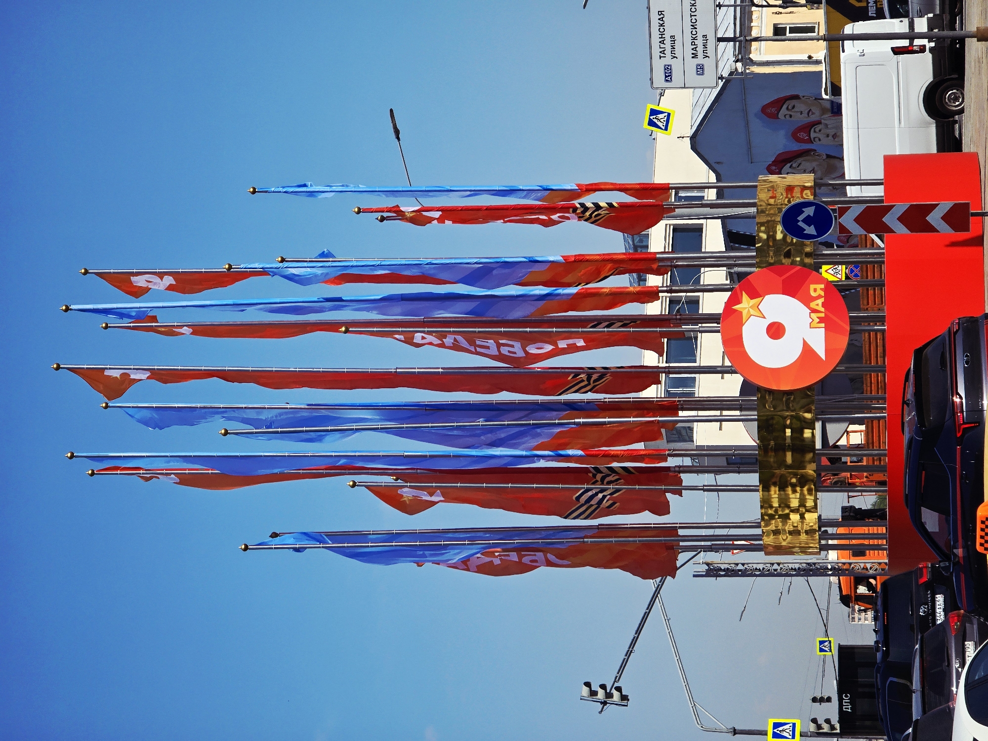 The image depicts a street in Moscow, Russia, decorated with flags and banners for the Victory Day celebration on May 9th. Numerous red and blue flags are prominently displayed on poles against a clear blue sky. A large sign with the number "9" and the word "MAY" in Russian is visible. In the background, there are buildings, a white van with a mural of women wearing red berets, and street signs indicating "Taganskaya Street" and "Marxists Street." Road signs, including pedestrian crossings and directional arrows, are also present. Several cars and taxis are parked along the street. The overall atmosphere suggests a festive occasion.
