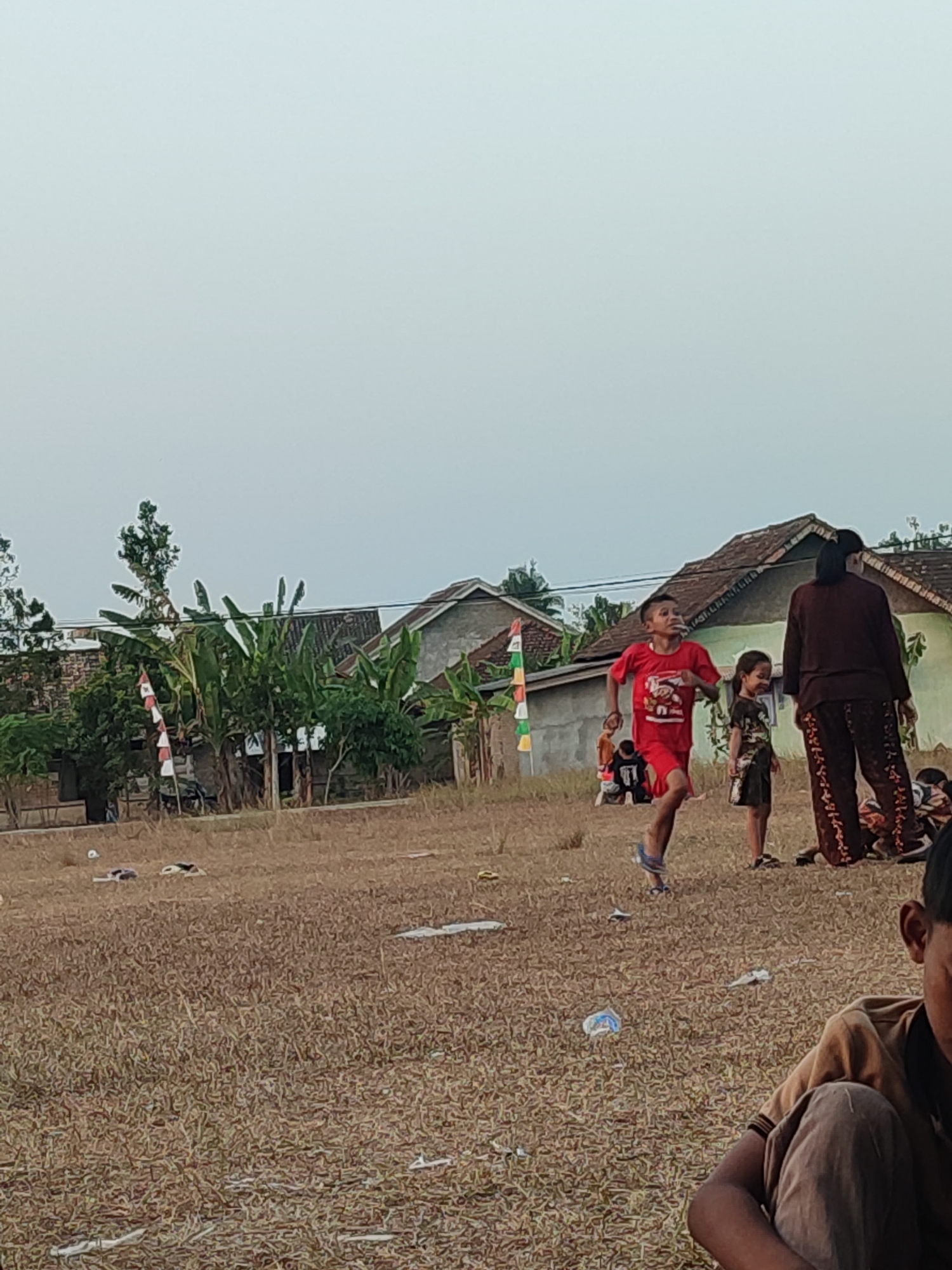 In a dry, grassy field in Nusa Jaya, Indonesia, several people are gathered near a cluster of houses. In the foreground, a young boy in a brown shirt and grey pants sits looking downwards. Further back, a boy in a red shirt and blue flip-flops is captured mid-stride, appearing to be running. He is accompanied by a younger girl in a patterned dress and a woman in a brown long-sleeved shirt and patterned pants. Several houses with tiled roofs are visible in the background, surrounded by banana trees and other vegetation. A flag with red and white stripes is planted near the houses. The ground is littered with various pieces of trash, including plastic wrappers and bottles. The sky is overcast and pale.