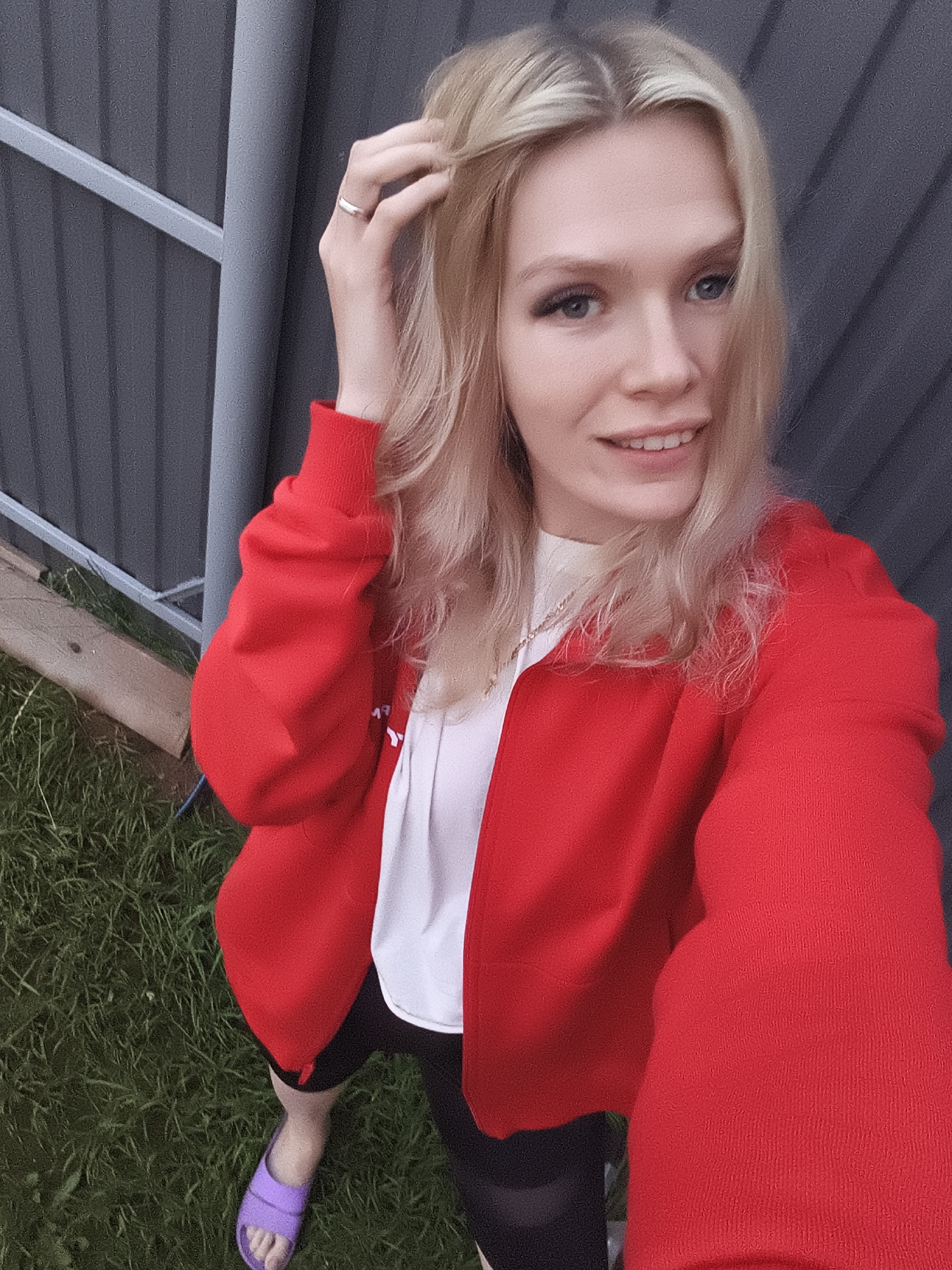 A young blonde woman is taking a selfie outdoors. She is wearing a red zippered jacket over a white t-shirt and black shorts. She has a silver ring on her right index finger and is touching her hair with that hand. Her left foot is visible, wearing a purple sandal. She is standing on grass, with a dark gray fence in the background. A wooden plank is visible on the ground near the fence.