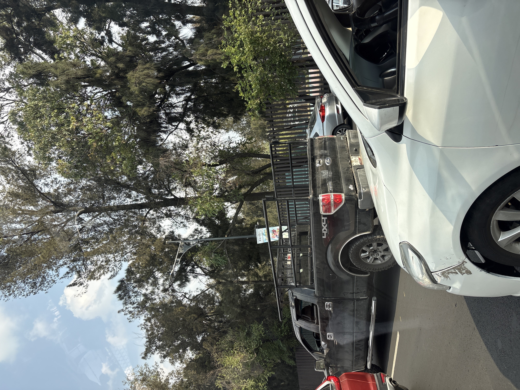 The image captures a street scene in Mexico City, Mexico. A white car with some damage on the front bumper is parked prominently in the foreground. Behind it, a black pickup truck with a metal rack is visible, followed by a silver car. A dark metal fence separates the street from a green space filled with trees. A street light with signage is mounted near the black pickup truck. The sky is partially visible through the trees, displaying a mix of blue and clouds.