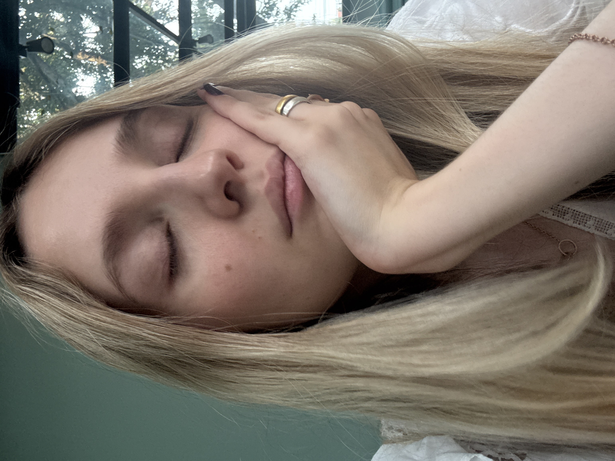 A close-up, tilted shot shows a fair-skinned woman with blonde hair resting her face on her hand. She has her eyes closed. She is wearing rings on her fingers and a bracelet. The setting appears to be indoors near a window, visible in the background. The image was likely taken in Mexico City, Mexico.