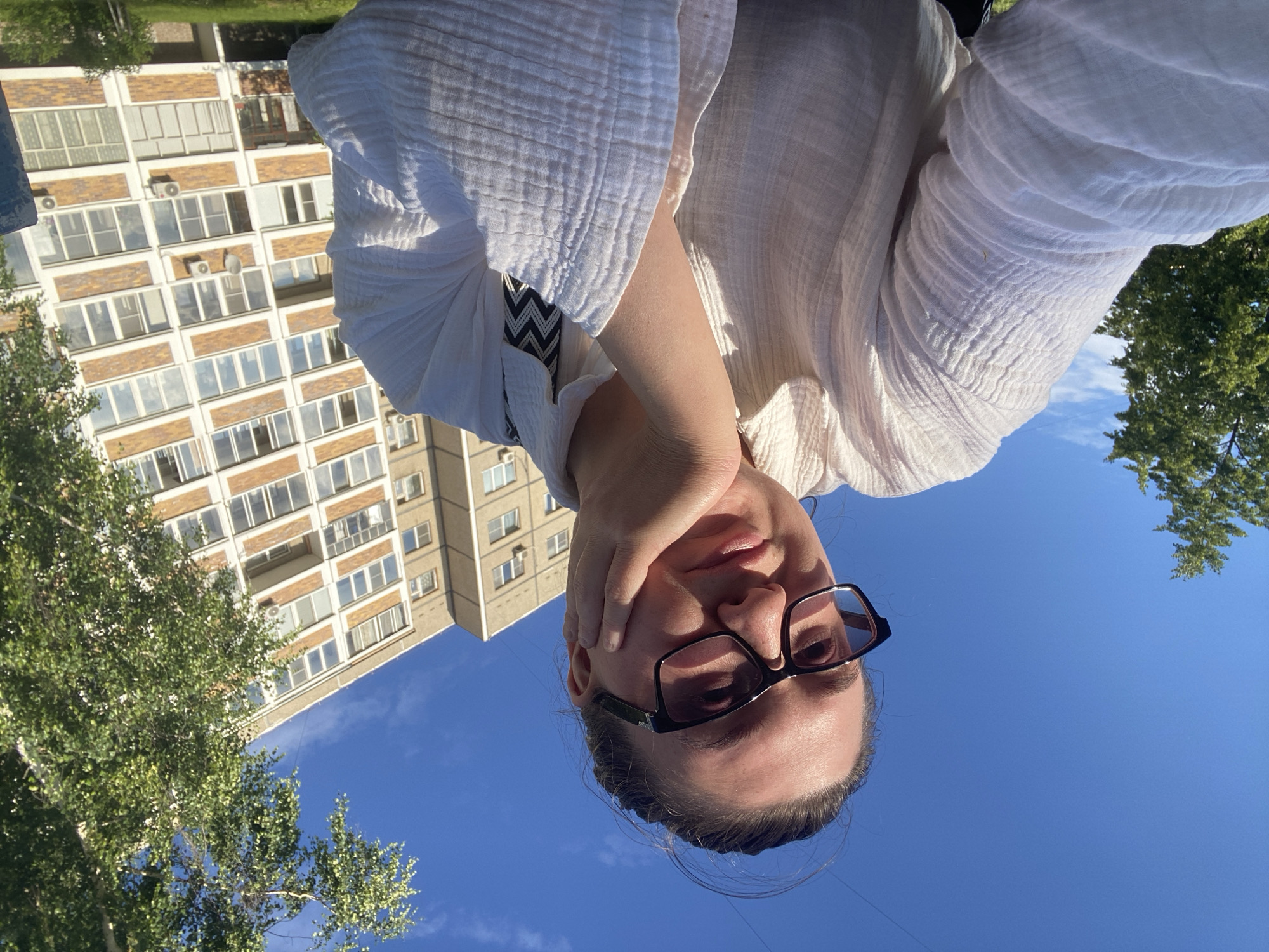 This is a selfie of a person with glasses, wearing a white crinkled fabric shirt, with their hand resting on their face. They are looking at the camera with a neutral expression. In the background, a multi-story building with many windows and balconies is visible, along with green trees and a clear blue sky. The sun appears to be shining, suggesting it is daytime. The image is oriented upside down. The image appears to have been taken in Chaikovsky, Russia, as indicated by the building style which is common in Russian apartment blocks, and the foliage suggesting a temperate climate.