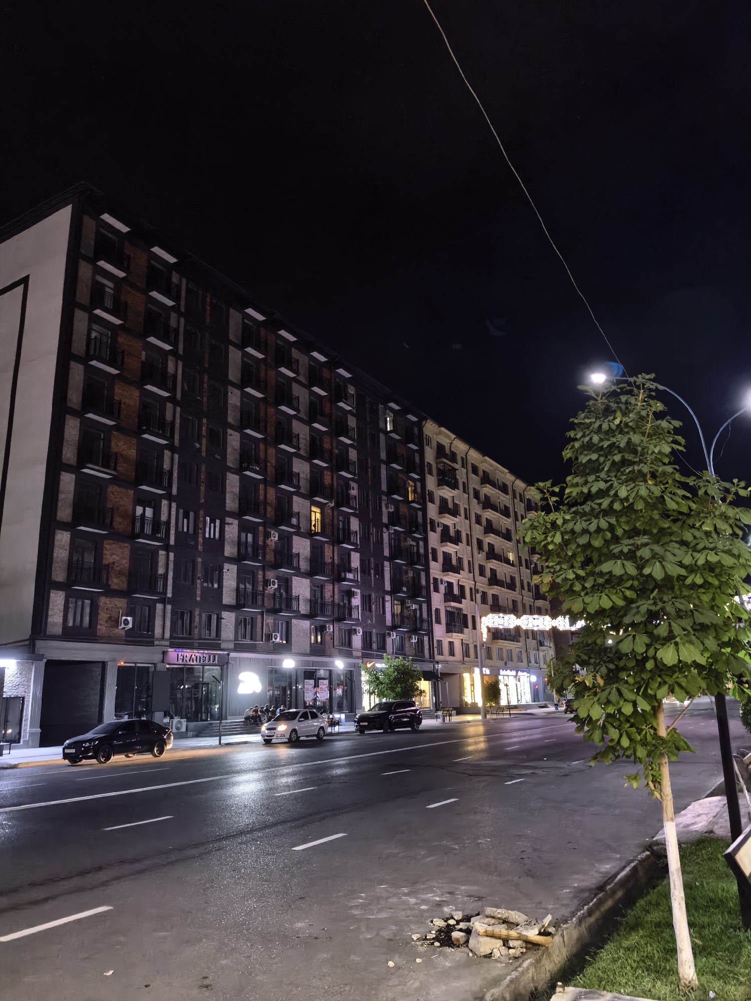 The image depicts a street scene at night in Fergana City, Uzbekistan. A multi-story apartment building with dark facade and brick accents dominates the left side of the frame. Its windows are illuminated, suggesting occupancy. To the right, a lighter-colored building stretches further down the street, also with visible lit windows. A wide, wet asphalt road occupies the foreground, marked with white lane dividers. Several cars are present on the road: a black sedan on the left, a white sedan in the middle lane, and a dark-colored vehicle further down the street on the right. A mature tree with lush green foliage stands on the right side of the sidewalk, illuminated by streetlights. The sidewalk itself appears to be made of concrete, with a curb bordering the road. Debris, including rocks and wood, is visible on the sidewalk near the curb. The sky above is dark and clear, indicating nighttime.