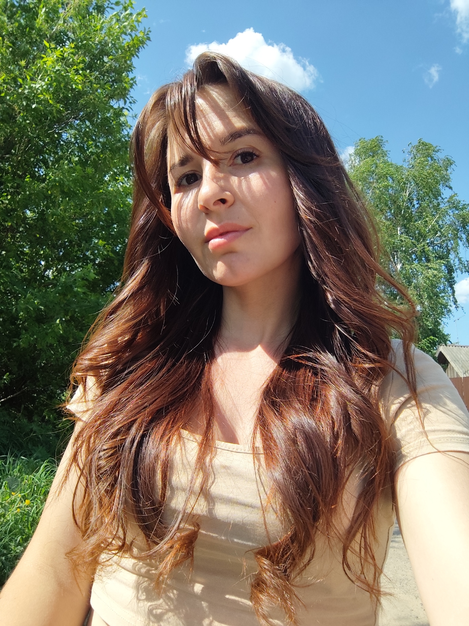 A woman with long, reddish-brown hair is posing for a selfie outdoors on a sunny day. She is wearing a light beige top. The background includes green trees and a blue sky with scattered clouds. A small section of a weathered building structure is visible in the background on the right. The exact location in the city of Фоки, Russia is not discernible from the image.