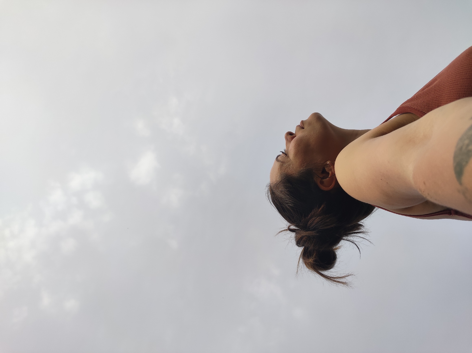 From a low angle, a woman with her head tilted back faces the sky. Her dark hair is pulled back into a messy bun. She wears a red tank top, and a tattoo is visible on her left arm. The background is a pale, overcast sky with some cloud cover. There is no indication of the location being in the city of Фоки, Russia.
