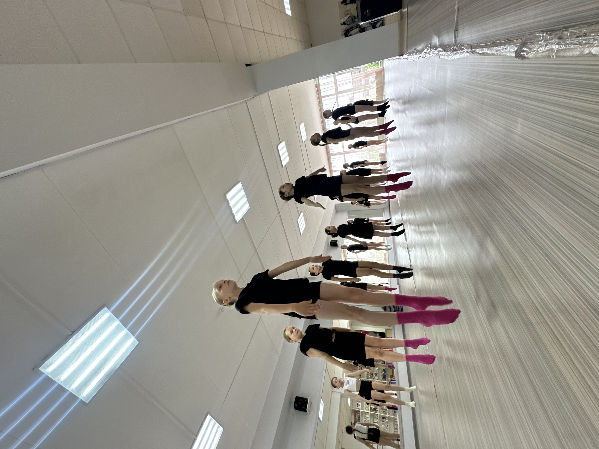 In a dance studio in Votkinsk, Russia, a group of young dancers in black leotards and shorts practice a routine. Most have pink socks on. They are lined up and standing with their arms extended, facing the wall, which is covered with a mirror. There is a set of lights on the ceiling, and shelves with items on the wall.