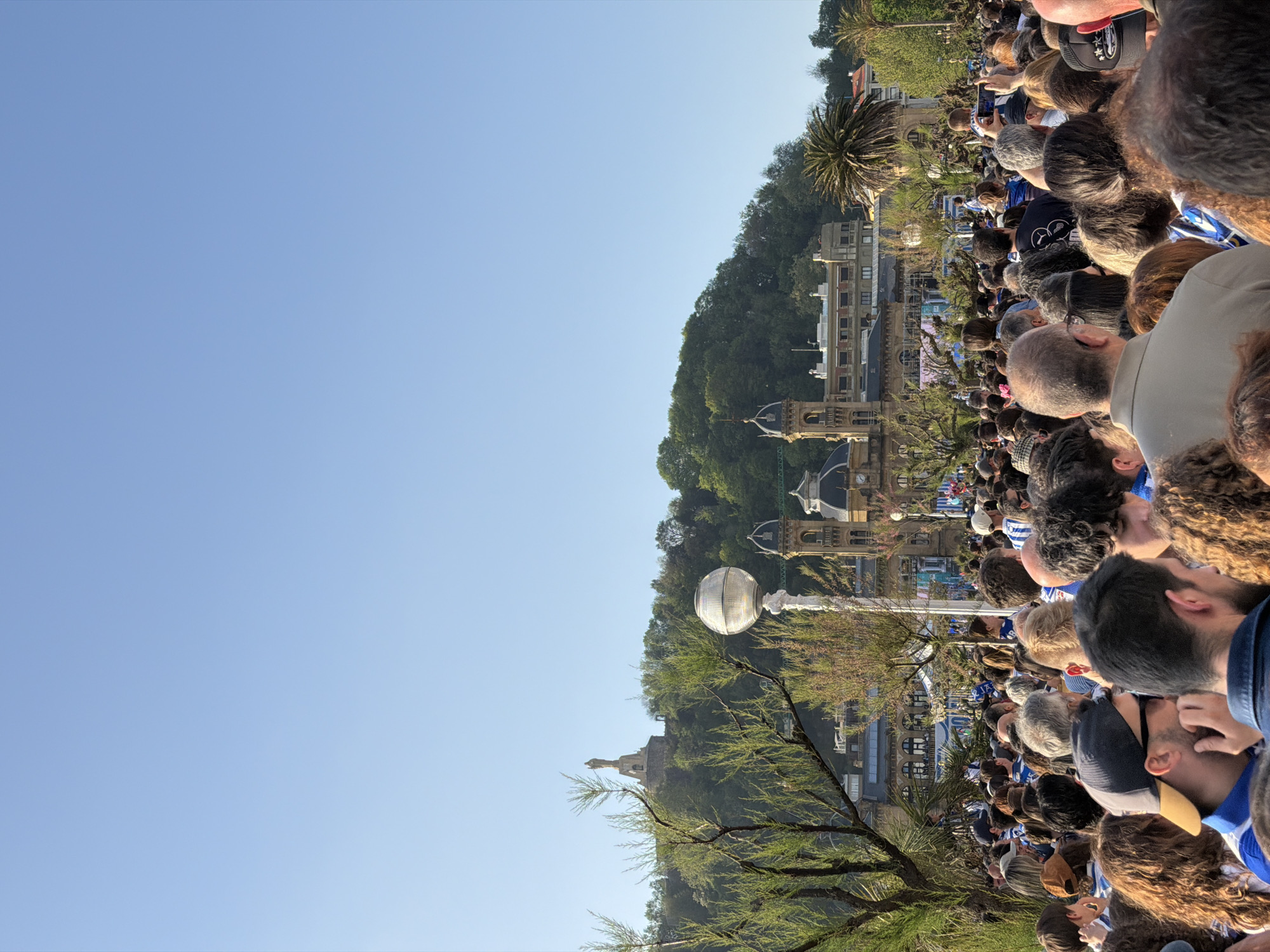 The image, when correctly oriented, depicts a large crowd gathered outdoors under a clear blue sky in San Sebastián, Spain. The foreground and midground are filled with the backs of numerous people, all facing towards a focal point beyond the left edge of the frame. Many attendees are dressed in blue and white striped attire, strongly suggesting support for a local sports team, likely Real Sociedad, known for these colors, during a celebratory event or parade. Some individuals are seen holding up their phones, presumably to capture photos or videos of the spectacle.
In the background, a picturesque urban setting unfolds. Elegant buildings with classical architectural details, including domes and ornate facades, line what appears to be a promenade or a street. Behind these structures, a steep, densely forested hillside rises, topped by a visible statue or monument, reminiscent of landmarks on Monte Urgull. A distinctive white, spherical streetlamp stands in the midground, partially framed by leafy trees.
The time of day is bright and sunny, indicated by the vibrant blue sky and the strong natural light illuminating the scene. The weather appears perfect for an outdoor gathering. The overall impression is one of a lively public event, with the crowd's focused attention and density conveying a sense of excitement and collective engagement. No legible text is visible on any signs or banners.