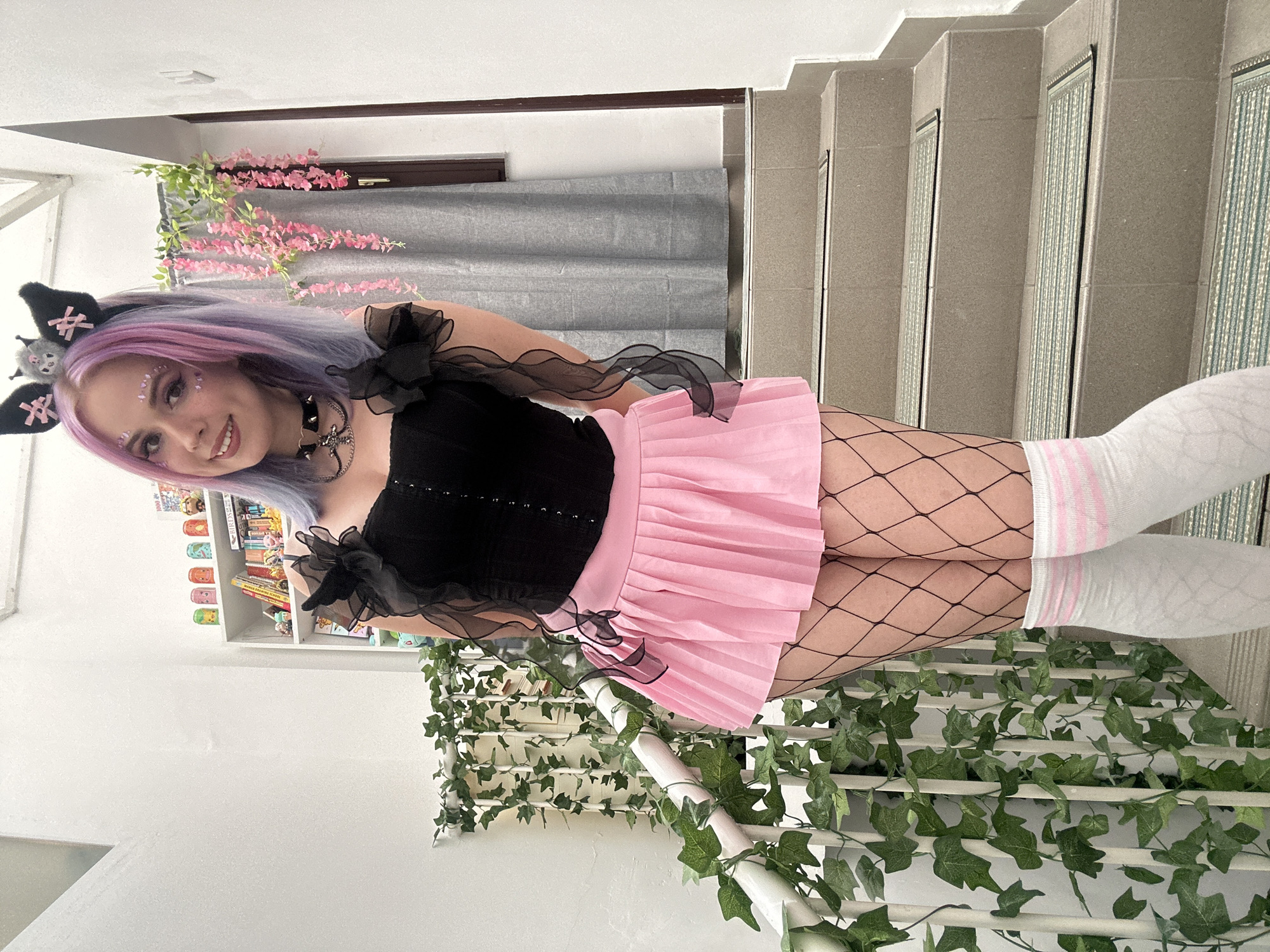 A young woman in a black corset top and pink pleated skirt with cat-ear headband and fishnet stockings is posing indoors, likely in a residential building. She is standing on stairs that have decorative strips of silver beads, which are part of the stair design. The stairs are white, adorned with artificial vines. She wears white knee-high socks with pink stripes, and black tulle decorates her top. Her hair is light purple. The background includes a window with a grey blind, artificial pink blossoms, and a white bookshelf. The image was taken in City of Zagreb, Croatia.