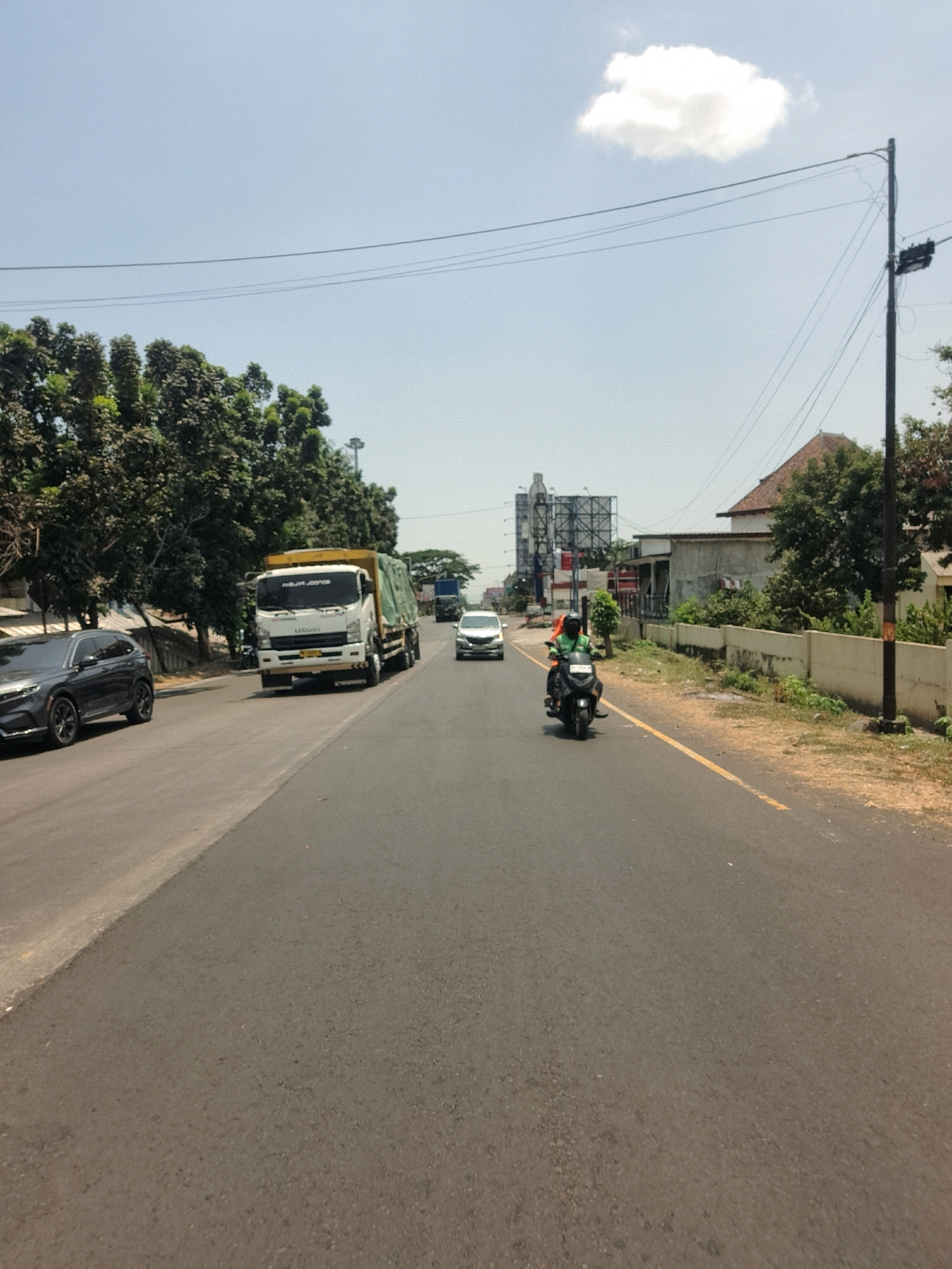 The image shows a paved road in Pandaan, Indonesia, under a clear blue sky with a single white cloud. A dark gray SUV is parked on the left side of the road, partially visible. Further down the road, a white truck with a green tarp covering its cargo is traveling away from the camera. A motorcycle with a rider wearing a green jacket and an orange helmet is also moving away from the camera. On the right side of the road, a concrete wall separates the road from a grassy area with trees and a building. Power lines are visible overhead, supported by a utility pole. In the distance, there are billboards and more buildings. The scene depicts a typical day on a road in Pandaan, with traffic and infrastructure elements.