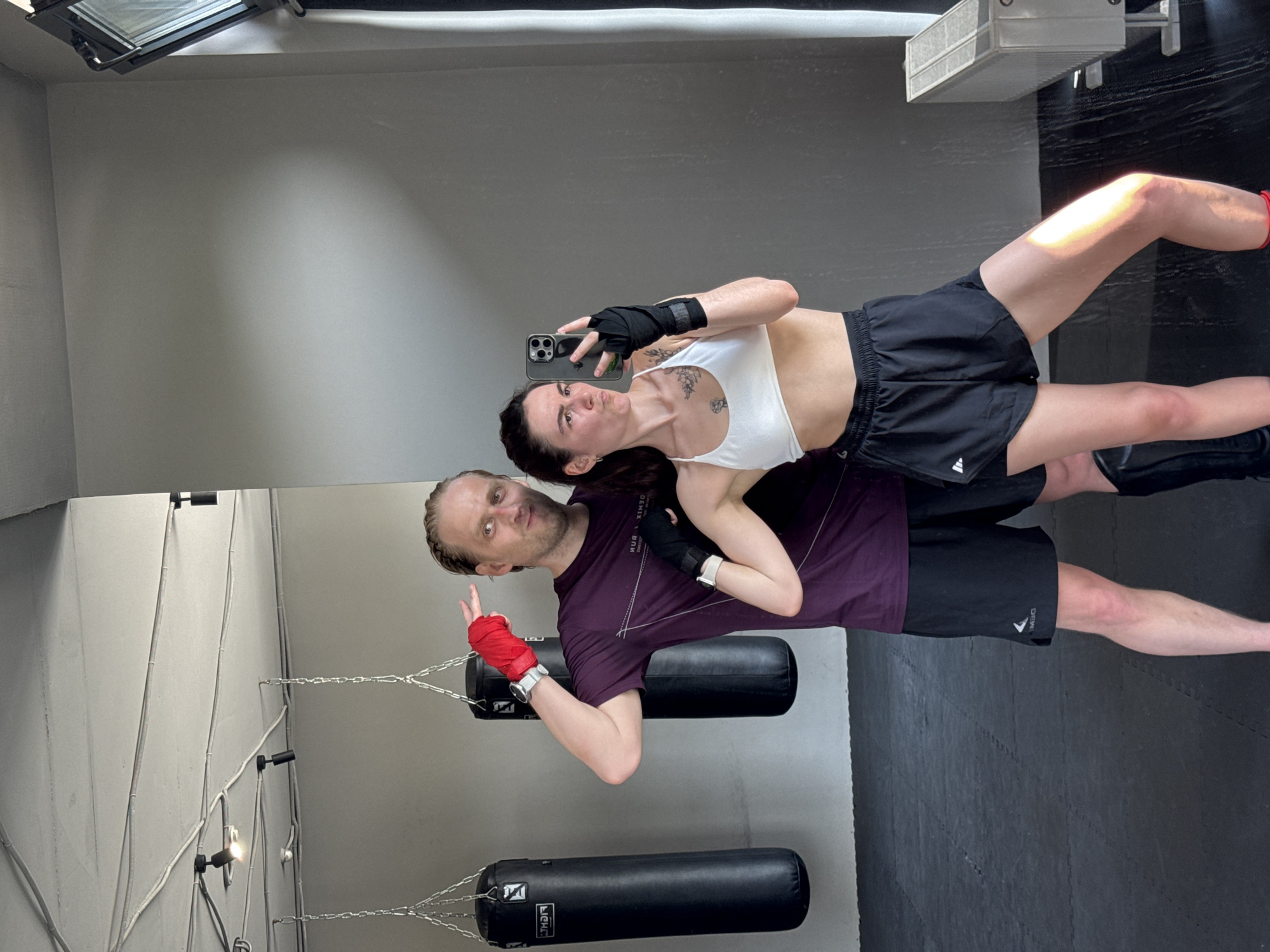 In a mirrored indoor boxing gym, two individuals, a man and a woman, pose for a selfie taken by the woman. Both are dressed in athletic wear. The woman wears a white sports bra, black shorts, and has black hand wraps. The man wears a purple t-shirt, black shorts, and has red hand wraps. Behind them are two hanging black punching bags. The location is presumed to be in Tyumen, Russia.