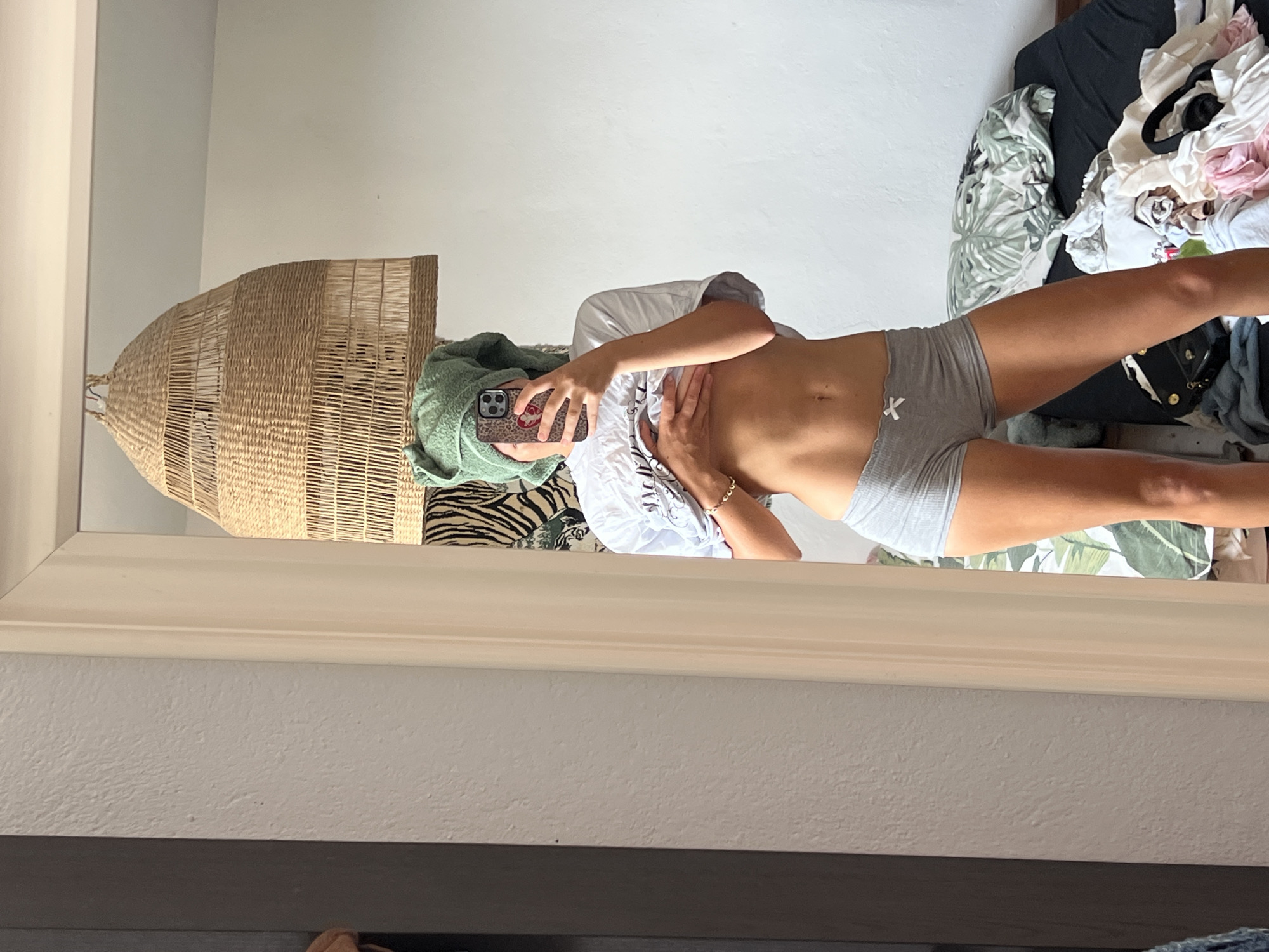 The image is a vertical, wide-angle, reflected shot in a mirror, showing a person and their surroundings. The person, who appears to be female, is wearing a white t-shirt and grey shorts. They are holding a phone with a leopard-print case in their right hand, while their left hand is on their stomach. A green towel is draped over their head.
In the background, a large, woven basket is visible, along with a pile of clothing and what appears to be bedding with a tropical leaf pattern. A dark surface, possibly a bed or dresser, is also visible. The lighting suggests it is daytime, with natural light entering the room.
The setting appears to be a bedroom or a dressing area. The overall mood is casual and personal, as indicated by the self-reflection and the casual attire. There is no visible text in the image. The location context of Mühldorf a. Inn, Germany, is provided but cannot be visually confirmed from the image itself.