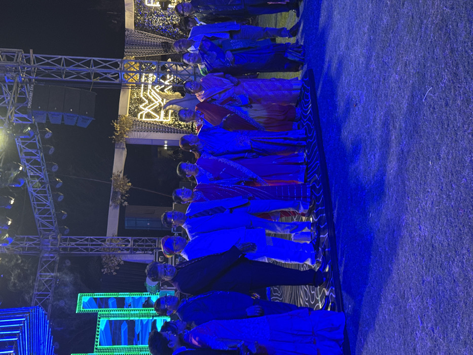 A group of individuals stand in a line on a patterned platform illuminated by blue and green lighting. Most are dressed in traditional Indian attire, including saris and kurtas. A stage structure with lights and sound equipment is visible in the background. The scene appears to be a performance or ceremony at an outdoor event.