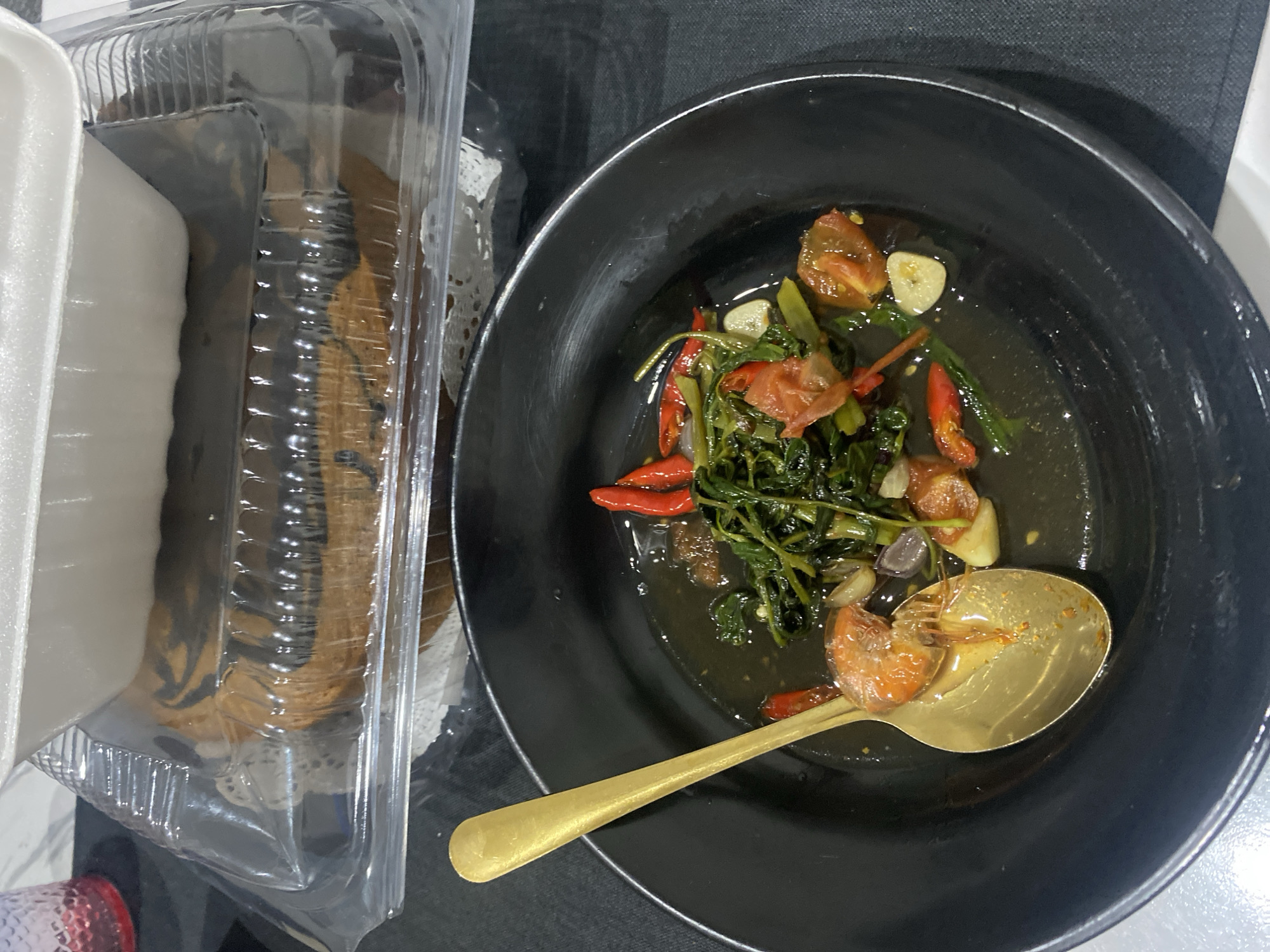 The image displays an overhead view of a meal setting indoors. In the foreground, a round, matte black bowl contains a dark-sauced dish. Visible ingredients in the bowl include vibrant green leafy vegetables, several whole red chili peppers, sliced white garlic cloves, and chopped red tomatoes. A gold-colored spoon rests partially submerged in the dish, holding a single cooked, orange-pink shrimp. The bowl is positioned on a dark gray or black fabric surface, which appears to be a placemat, with a white surface visible to the lower right.
In the background, two stacked food containers are present. The top container is a white, opaque foam or plastic takeout box lid, partially obscuring the item below. Beneath it, a clear plastic clamshell container holds a round, baked good, possibly a cake or bread, featuring a distinct dark brown swirl pattern on a lighter brown base. This baked good rests on a white, lacy paper doily. A translucent red cup or glass is partially visible on the far left in the background. The specific location of the scene in the city of Cibinong, Indonesia, cannot be visually confirmed from the provided image.