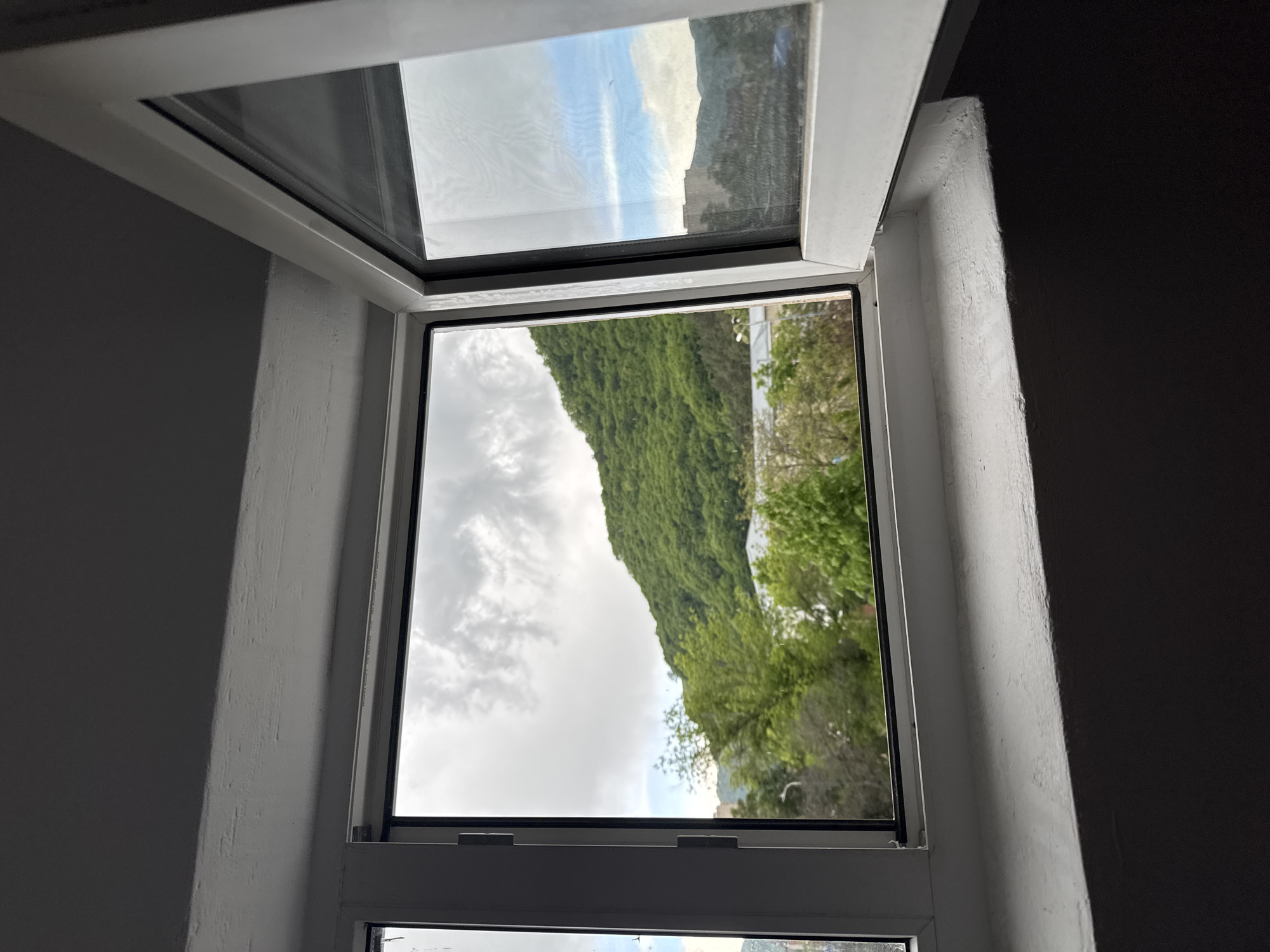 A view from inside a room looking out of an open window. The window has a white frame, and it appears to be an open-outward style window. Outside, a hillside covered in lush green vegetation and some trees is visible. The sky is overcast with clouds. The surrounding walls of the room are painted white and grey. There is no discernible evidence indicating the location of Karachayevsk, Russia.