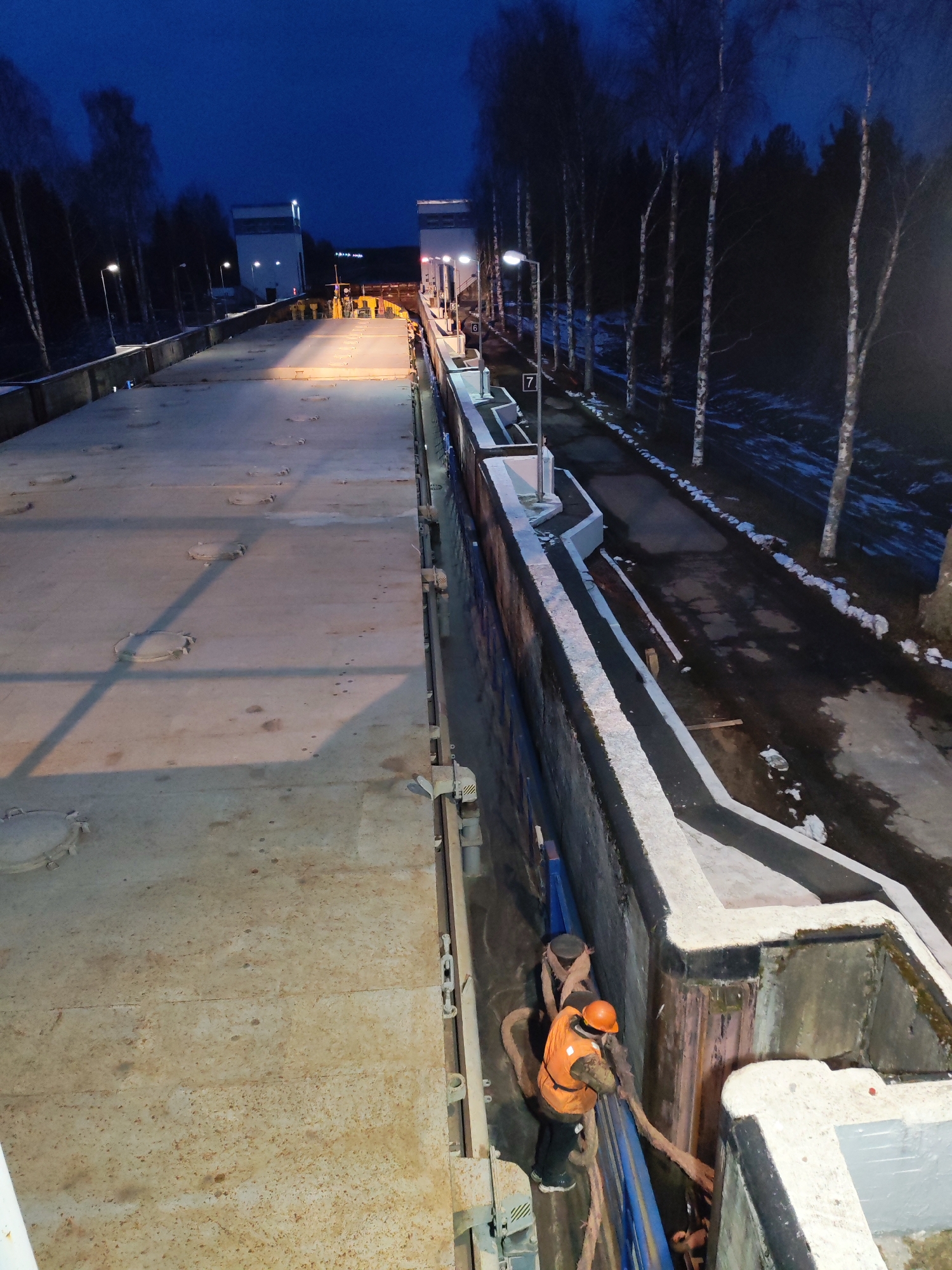 The image captures an industrial maritime scene at dusk or night, likely at a canal lock or a large quay. A spacious, flat-decked vessel, possibly a barge, dominates the left side of the frame, its surface appearing weathered with rusty patches and featuring numerous circular access hatches.
On the right, a worker in an orange safety vest and helmet is actively engaged near the concrete wall of what appears to be a lock chamber. They are handling thick, textured ropes or hoses, suggesting tasks related to mooring, pumping, or operational adjustments. The concrete structure extends into the distance, bordered by a dark path and a line of tall, slender, bare trees, resembling birches. Patches of snow are visible on the ground beside the trees, indicating cold weather.
Artificial lights from streetlamps illuminate the scene, casting shadows and highlighting details against the deep blue twilight sky. In the background, two distant, tall buildings or control towers are visible, their upper sections lit. A yellow vehicle can be faintly seen between them. Visible text includes the numbers "6" and "7" on white signs attached to the concrete walls. The overall ambiance is functional and quiet, under the glow of artificial lighting.