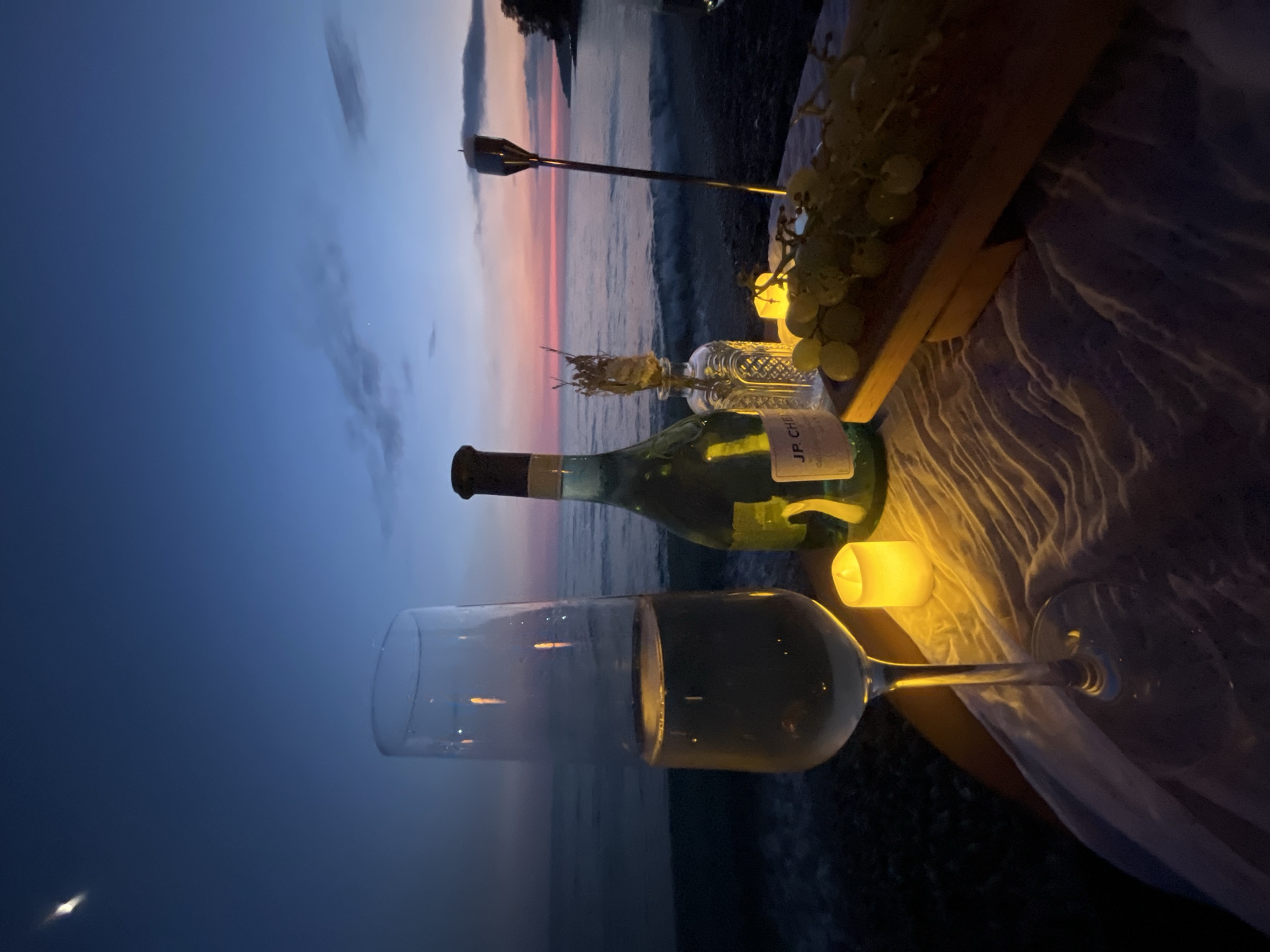 A table is set for a romantic evening on a pebble beach at dusk. A bottle of champagne, a flute filled with champagne, a bunch of grapes on a wooden board, a lit candle, and a decorative bottle with dried flowers are arranged on a white cloth. In the background, the Black Sea stretches out towards the horizon, with gentle waves lapping the shore. The sky is a gradient of deep blue, transitioning to hues of pink and orange as the sun sets. The location is the city of Sochi, Russia. There are no people present in the image.