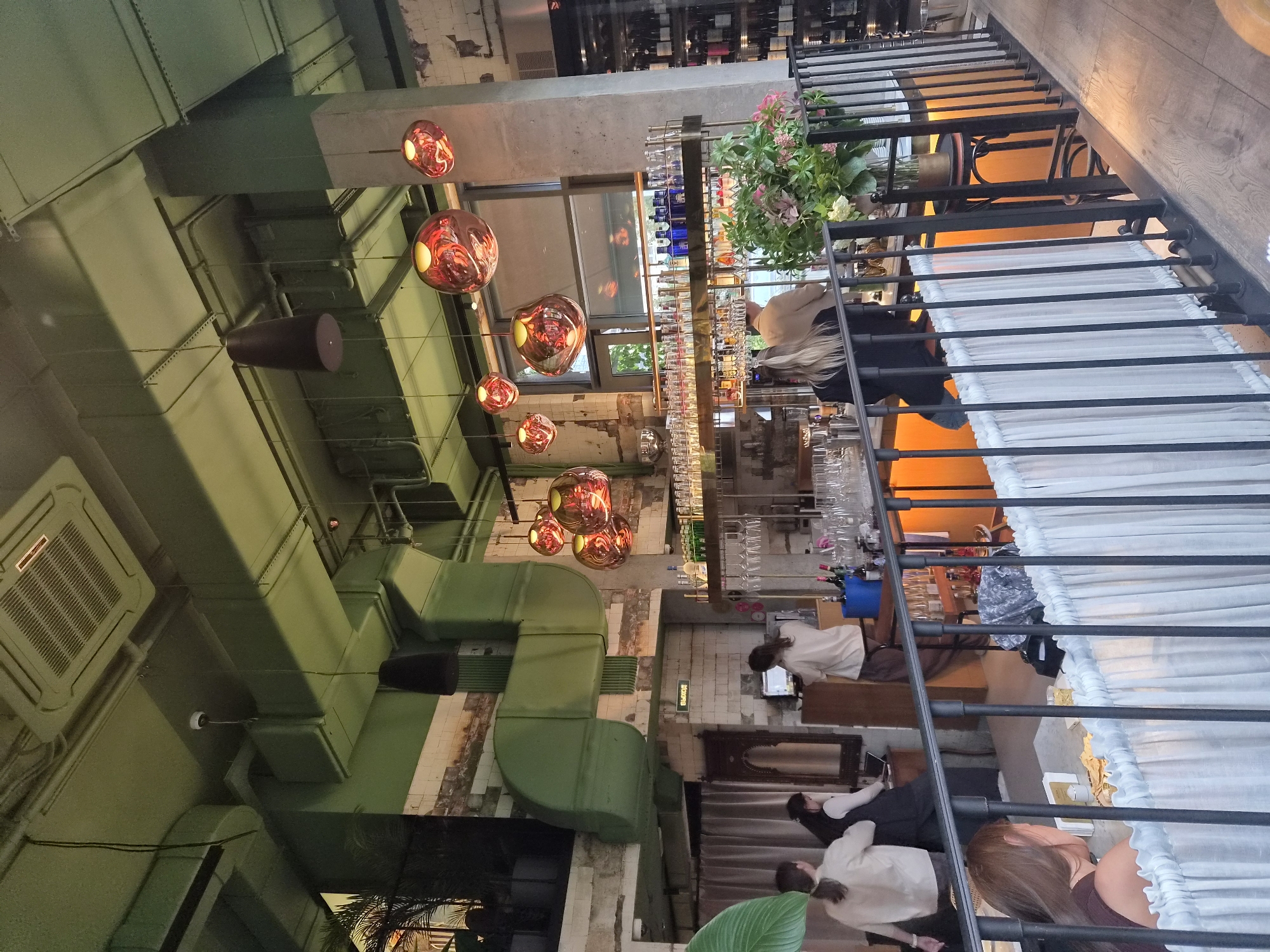 The image captures an interior view of a modern restaurant or bar in Saint Petersburg, Russia. The space features an industrial aesthetic with exposed green-painted ductwork and light-colored tiled walls. Hanging from the high ceiling are several artistic, bulbous pendant lights with a metallic, reflective surface, as well as a darker, conical pendant. The main area visible includes a bar with a bright orange countertop, stocked with glasses and bottles. Several individuals are present in the scene. Two women are standing behind the bar, one appearing to be working at a computer. Further to the left, three individuals, two women and one person whose gender is unclear, are interacting near a curtained area. In the foreground, a woman with brown hair is seated at a table, partially obscured by a white, pleated fabric barrier and a black metal railing. The overall atmosphere suggests a contemporary dining or drinking establishment.
