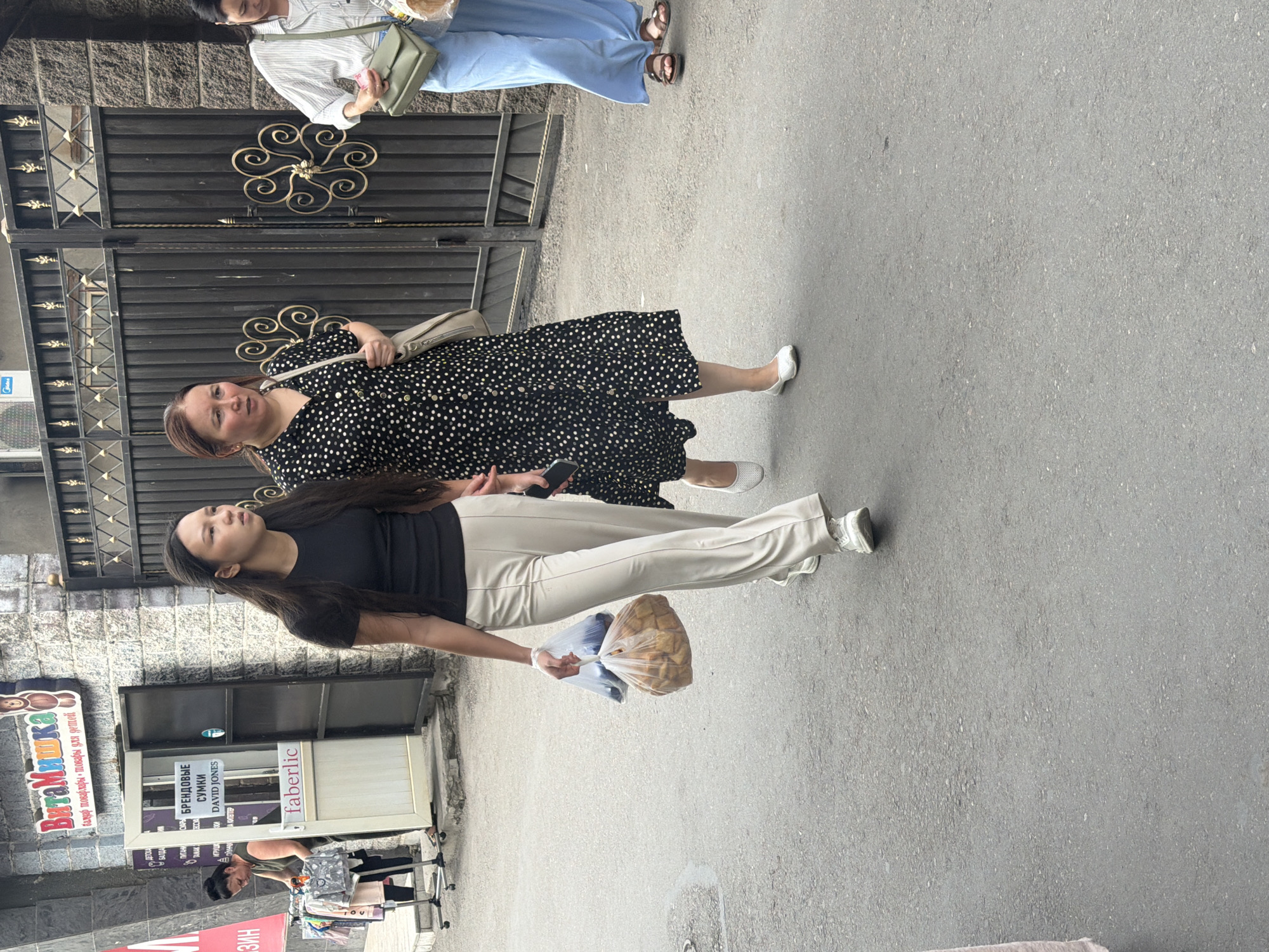Two women are walking on an asphalt street in front of a building. The woman on the left is younger, with long dark hair, wearing a black t-shirt and light-colored pants. She is carrying two plastic bags filled with food. The woman on the right is older, with brown hair, wearing a black dress with white polka dots. She is carrying a light-colored shoulder bag. In the background, there is a building with signage, a black metal gate, and a clothing rack with children's clothes displayed. Another woman in a striped shirt and blue pants is partially visible on the far right, walking away from the camera. The scene appears to be in a commercial or market area.