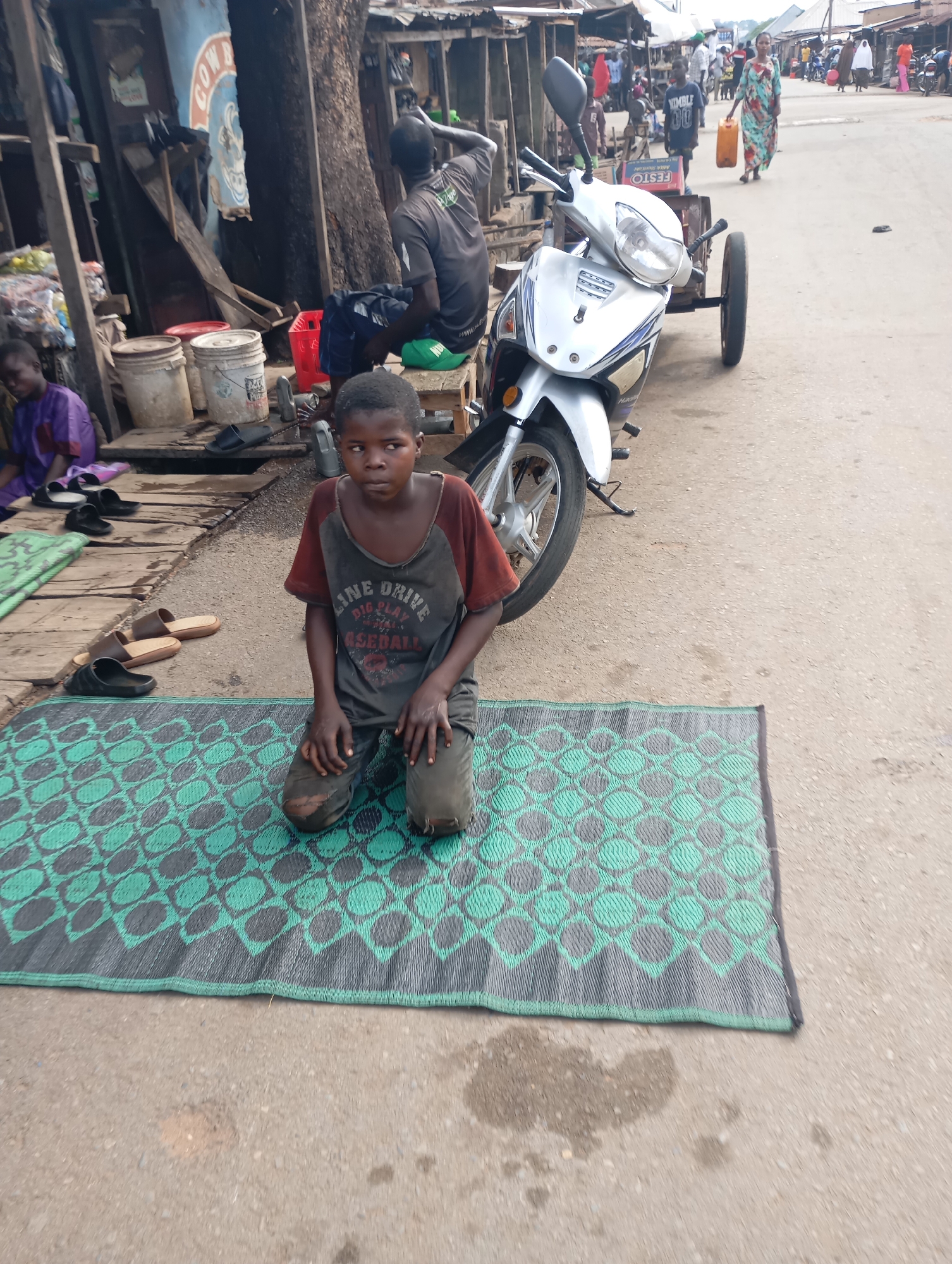 In the city of Shendam, Nigeria, a young boy kneels on a green and grey mat in a dusty street. He wears a worn, dark t-shirt with faded lettering and tattered pants. Beside him, a silver motorcycle with a trailer attached is parked on the road. In the background, various market stalls, some constructed from corrugated metal, line the street. A few individuals are visible in the distance, including a woman carrying a yellow container. The scene depicts a bustling street market environment.
