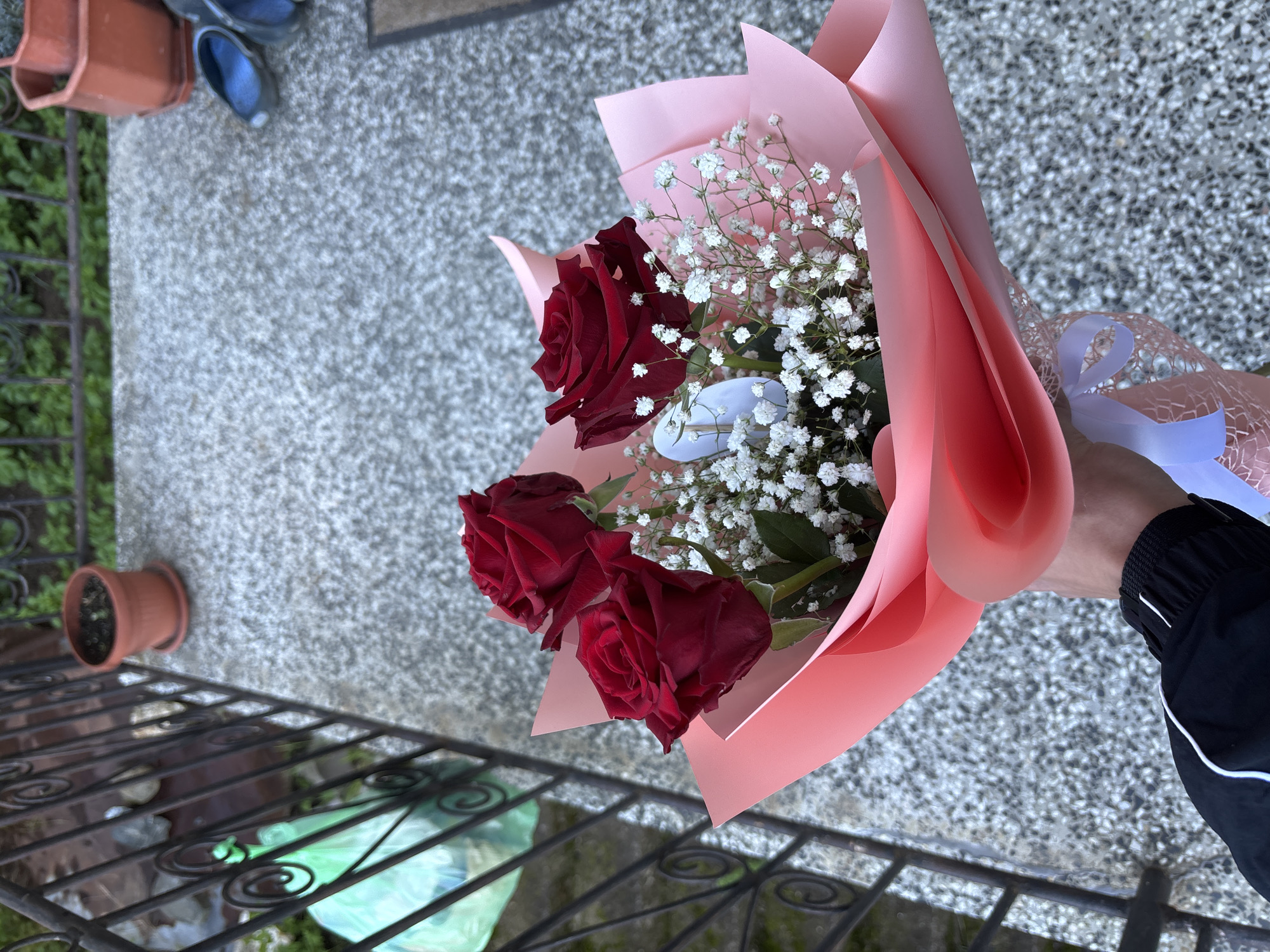 The image shows a hand holding a bouquet of three red roses and baby's breath flowers wrapped in pink paper and tied with a white ribbon. The person wearing a black jacket with a white stripe on the sleeve. In the background, there is a gray concrete surface, a black metal railing, a small brown flower pot, and a pair of blue slippers. Based on visual features such as building materials and landscaping style, the location may be the city of Zlatograd, Bulgaria.