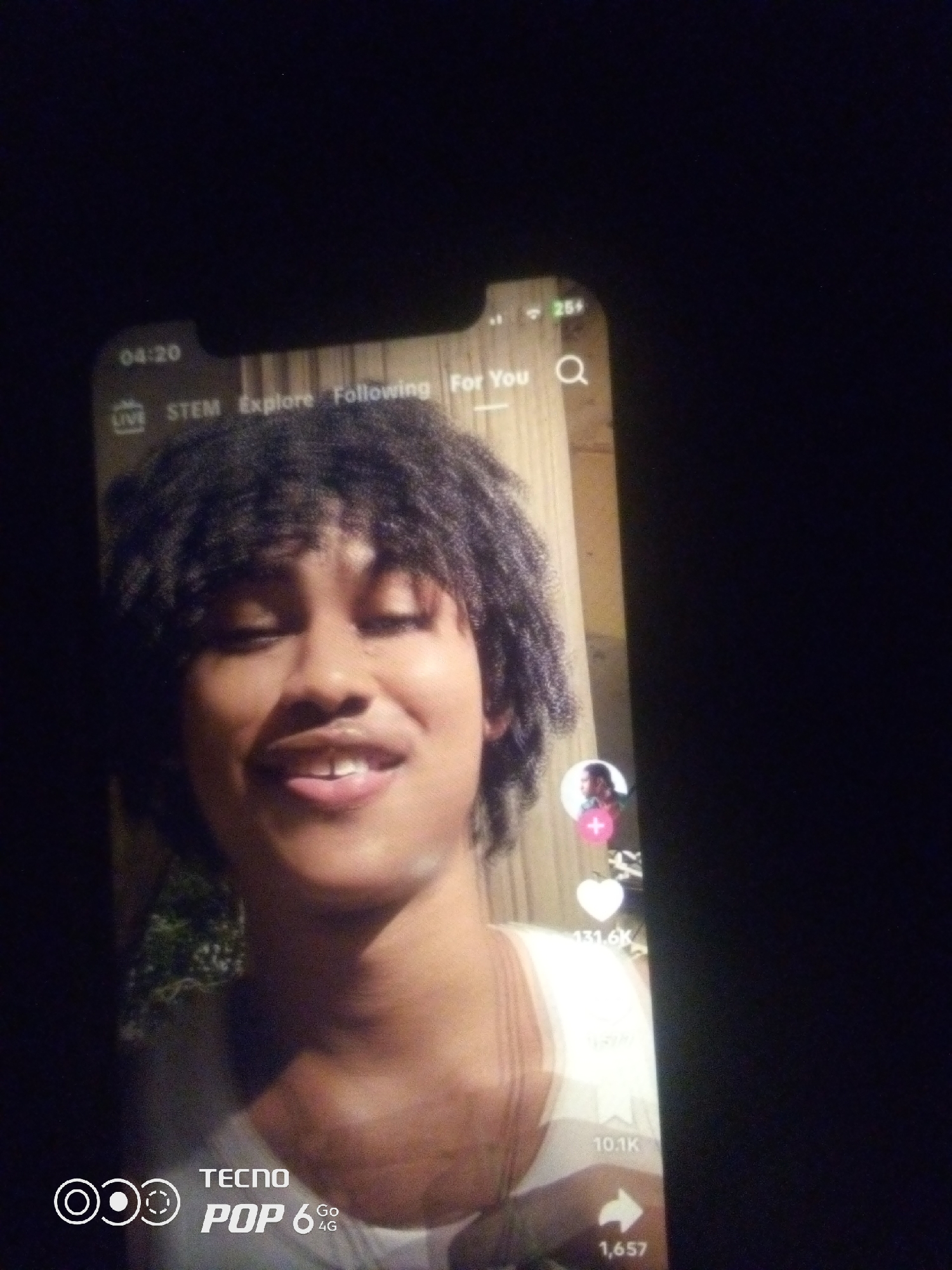 A close-up, low-light, vertical photograph captures a young person with dark, curly hair smiling directly at the camera. The person is wearing a white tank top. The image appears to be a screenshot from a mobile phone, with the phone's screen interface visible at the top, including the time "04:20" and menu options like "LIVE," "STEM," "Explore," "Following," and "For You." To the right of the screen interface, a battery icon and a signal strength icon are visible. On the right side of the image, within the phone screen, a circular profile picture of another person with a pink plus sign is superimposed. Below this, icons for likes, comments, and shares are visible, with numbers indicating "131.6K," "1,657," and "10.1K" respectively. The bottom left corner of the phone screen displays "TECNO POP 6 4G" with a circular logo. The overall lighting is dim, with the subject's face being the most illuminated area. No specific location in Howayi, Nigeria, is identifiable from the visual information in the image.