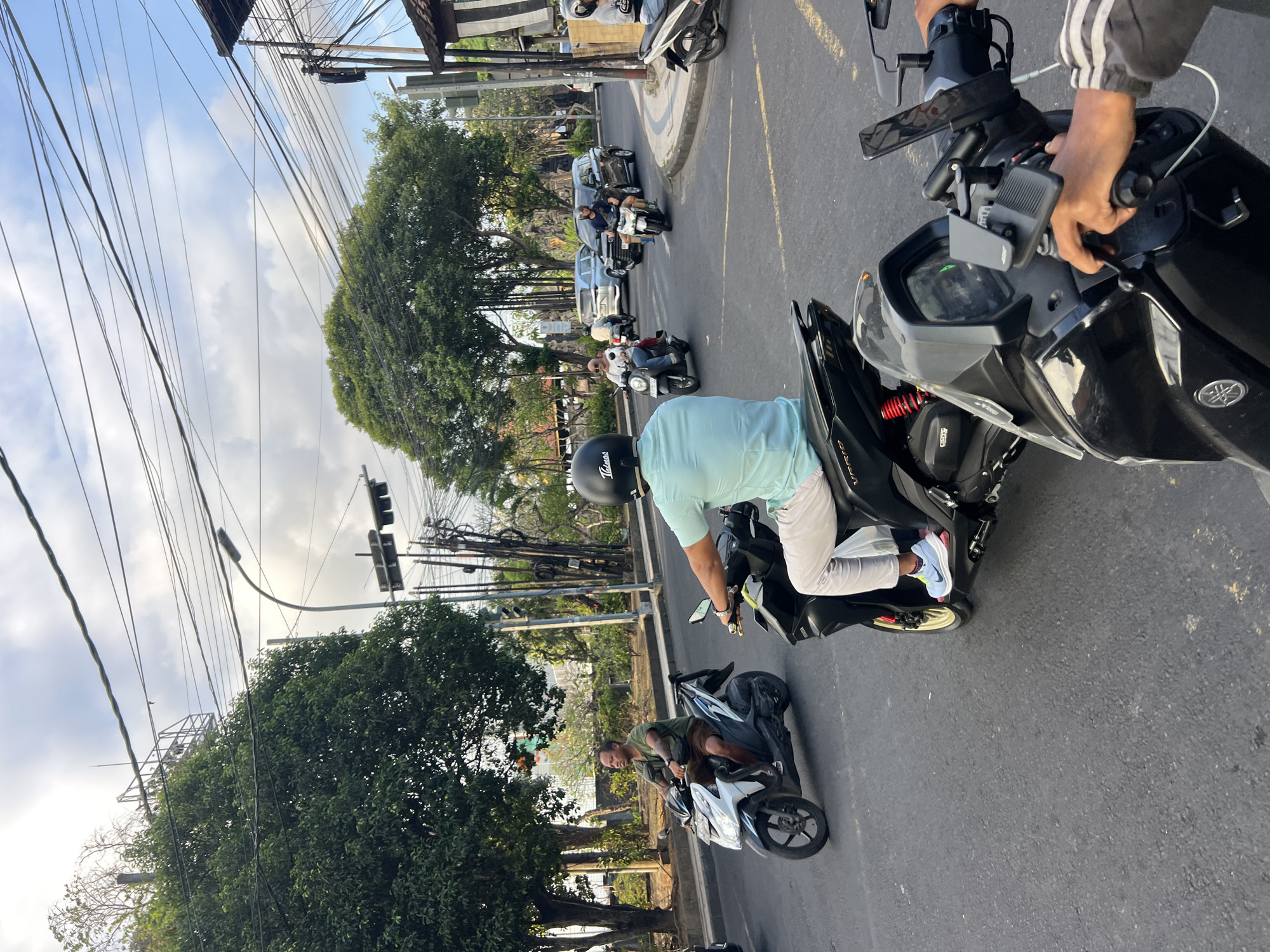 The image depicts a street scene in Kuta, Indonesia, characterized by traffic and a busy intersection. Multiple motorbikes are in motion, with riders navigating the road. One rider in the foreground is wearing a light blue shirt, white pants, and a black helmet, astride a black scooter. To his left, another man on a white and blue scooter wears a green t-shirt and shorts. In the background, several other motorbikes and cars are visible, including a white SUV and a black SUV. The environment includes mature trees lining the street, overhead electrical wires, and traffic lights. The sky is partly cloudy, suggesting a late afternoon or early evening setting. The overall impression is one of typical urban traffic flow in a tropical location.