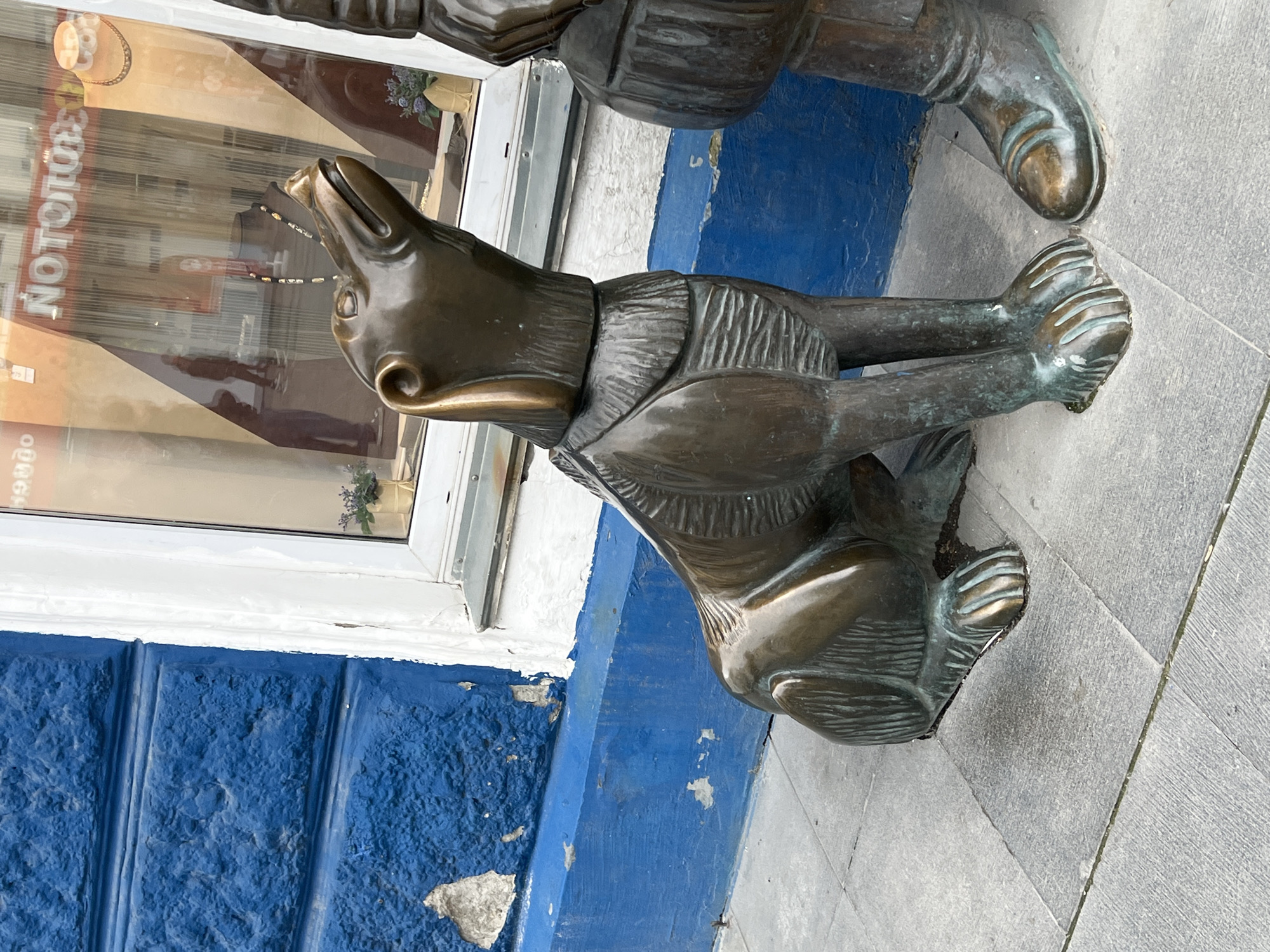 A bronze sculpture of a dog sits on a sidewalk in front of a building with a bright blue, textured facade. The dog is positioned in a seated posture, looking upwards with its mouth slightly open. Its fur is rendered with vertical lines, and it wears a collar-like detail around its neck. To the right of the dog, the leg and foot of another bronze sculpture, presumably a human, are visible. The human sculpture wears a boot. The sidewalk is paved with gray rectangular tiles. Behind the sculptures, a storefront window reflects the street and pedestrians. The building features a white-trimmed window frame and a blue painted base. The text "NOTOPOGON" is visible on an orange banner inside the storefront. The scene appears to be located in Yekaterinburg, Russia.