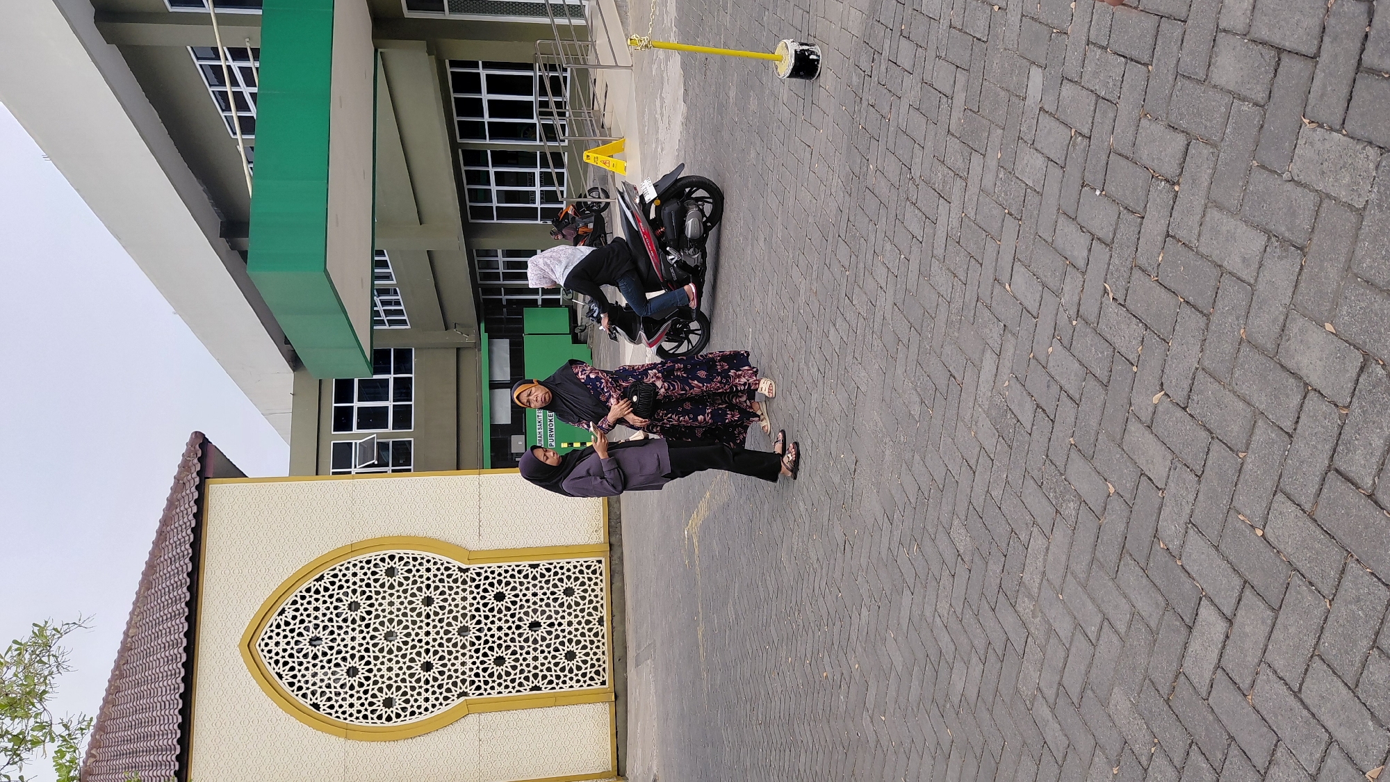 The image depicts an outdoor scene in the city of Rejasari, Indonesia, likely a parking lot or courtyard of a building. In the foreground, a grey brick pavement is visible. Two women are standing and conversing on the pavement. The woman on the left wears a dark hijab and a grey long-sleeved top with black pants. The woman on the right wears a black hijab and a dark floral-patterned dress. Behind them, a woman on a red and black scooter is positioned. She wears a light-colored hijab and blue jeans. The background features buildings with a blend of architectural styles. To the left, a cream-colored building with an arched window featuring intricate geometric patterns is prominent. To the right, a larger building with a dark green facade and numerous windows is visible. A bright green overhang extends from this building. The sky is overcast. A yellow pole with a black base and a yellow chain is situated in the mid-ground.
