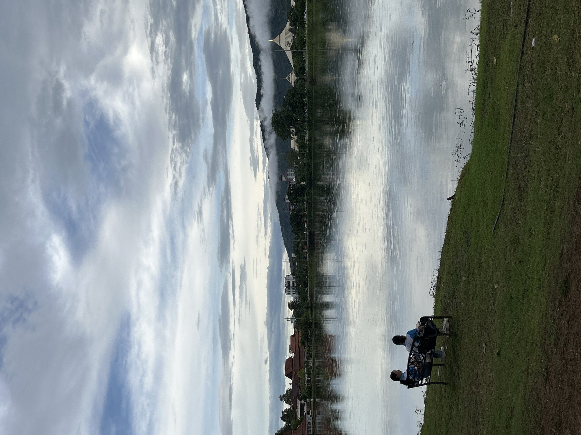 In Chiang Mai City Municipality, Thailand, two individuals are seated on a dark-colored bench facing a large body of water under a cloudy sky. The bench is positioned on a grassy incline overlooking the water. The person on the left, with dark hair, is wearing a blue and white patterned shirt and dark pants. The person on the right, also with dark hair, is wearing a blue t-shirt and dark pants. The water reflects the sky and the surrounding landscape, with ripples visible on its surface, suggesting it might be raining. Across the water, a cityscape is visible, featuring buildings with red roofs, modern multi-story structures, and lush green trees. In the distance, a mountain range is shrouded in clouds, with a distinctive golden-domed building prominent against the misty backdrop. The overall atmosphere is serene and slightly overcast.