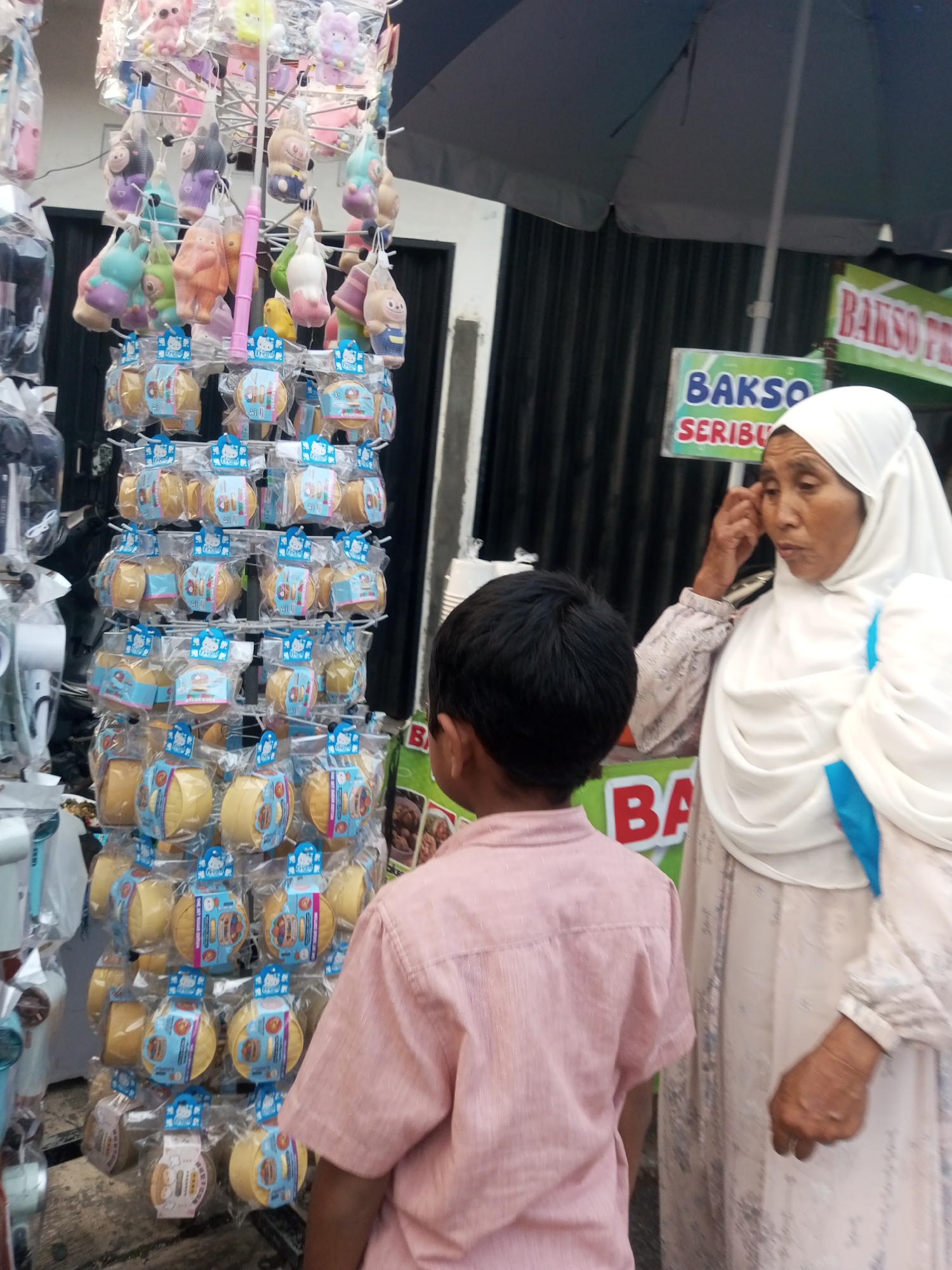 This is an outdoor medium shot of a small child and an adult woman standing at a food stall. The child, viewed from the back, wears a pink collared shirt. The woman, wearing a white hijab and a light-colored patterned dress, stands to the child's right and looks to her right, her right hand touching her temple. Behind them is a rack of packaged toys and food signs. The food stall appears to be on a street in Pandansari, Indonesia. The ground is paved, and there is a corrugated metal wall in the background.